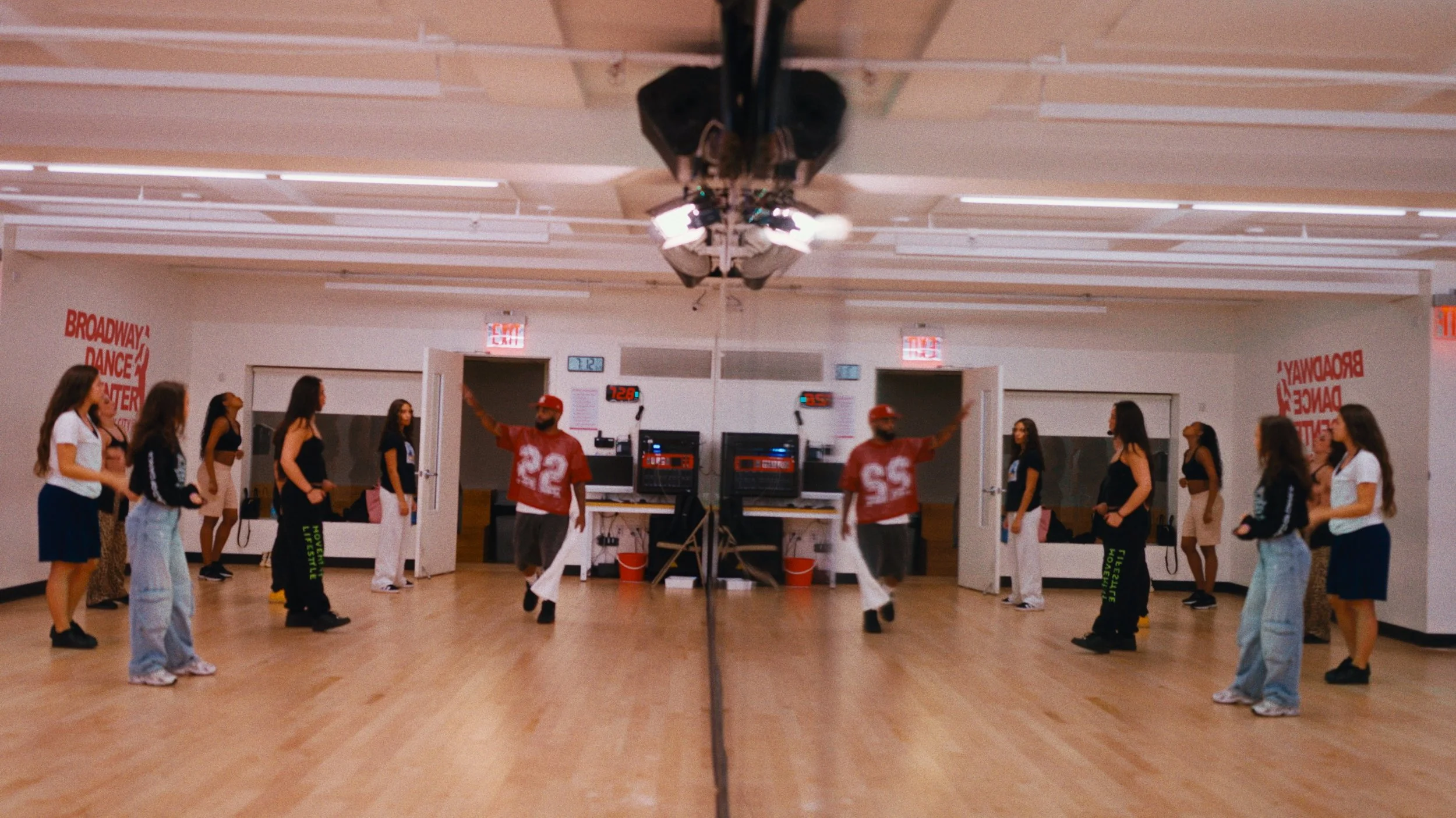 Dance rehearsal in a studio with people in two lines and two dancers in red jerseys practicing in the center.