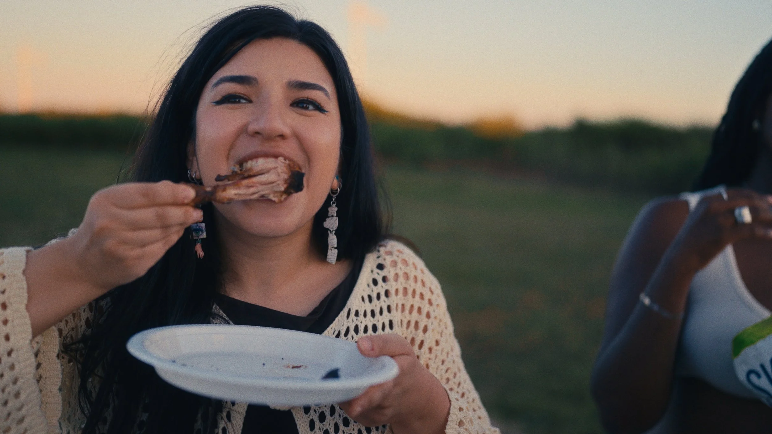 Woman with long dark hair eating a piece of grilled meat with a fork outdoors during sunset, holding a white plate.