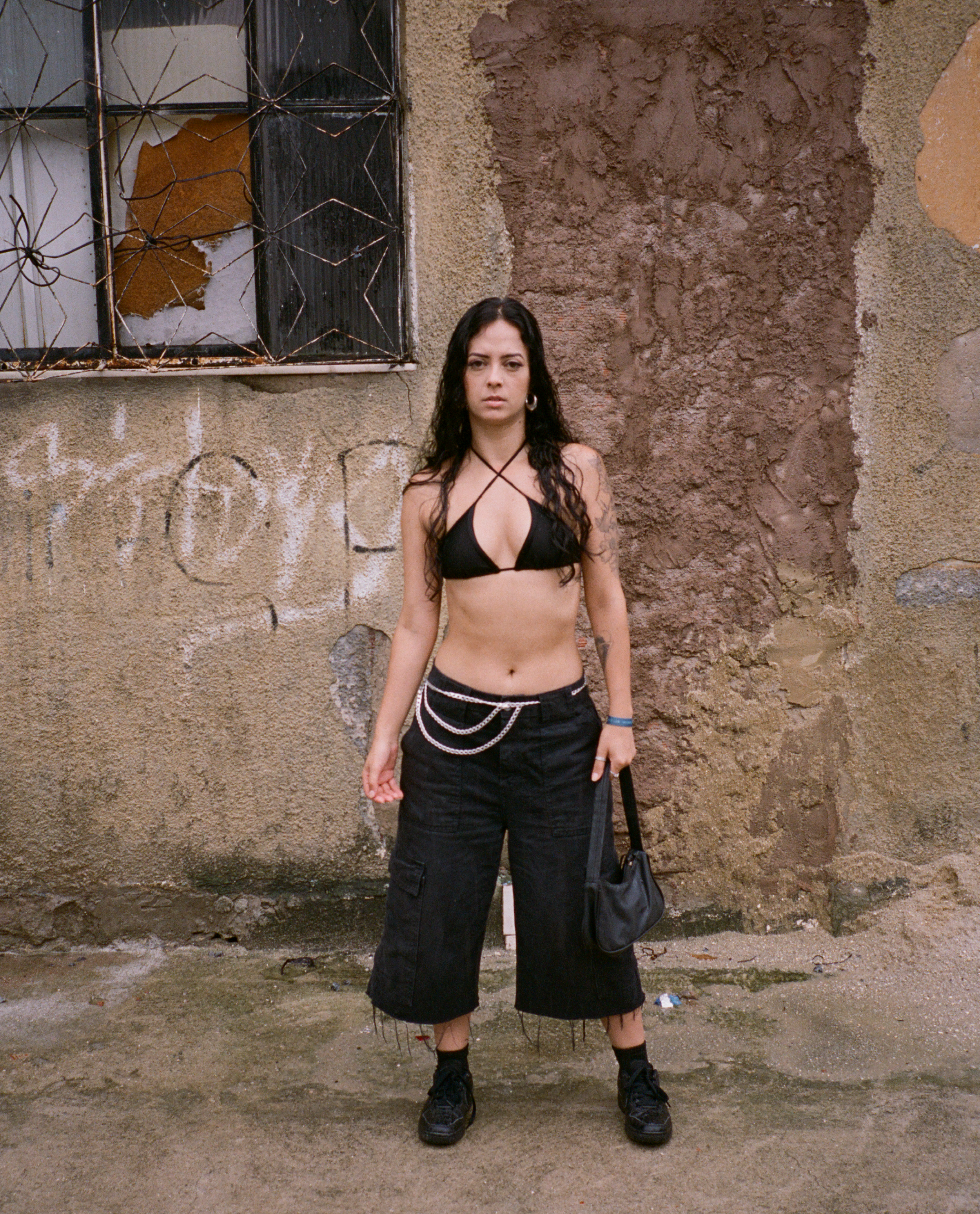 A young woman with long dark curly hair, wearing a black bikini top, black cropped cargo pants with chains, black sneakers, and carrying a black bag, standing against a weathered, graffiti-marked wall with broken windows.