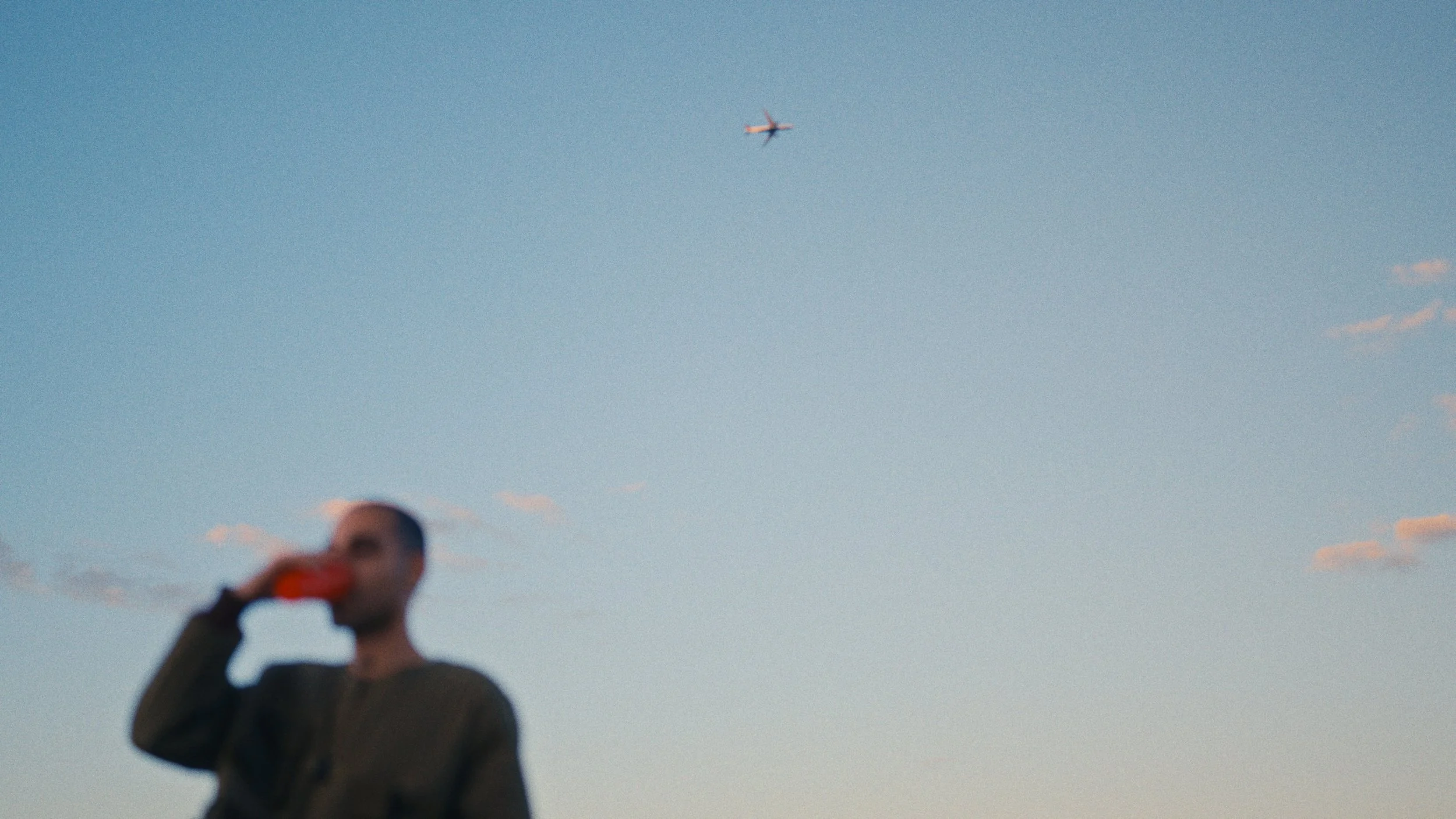 Blurred person with short hair drinking from a cup or bottle against the sky, with an airplane flying overhead.