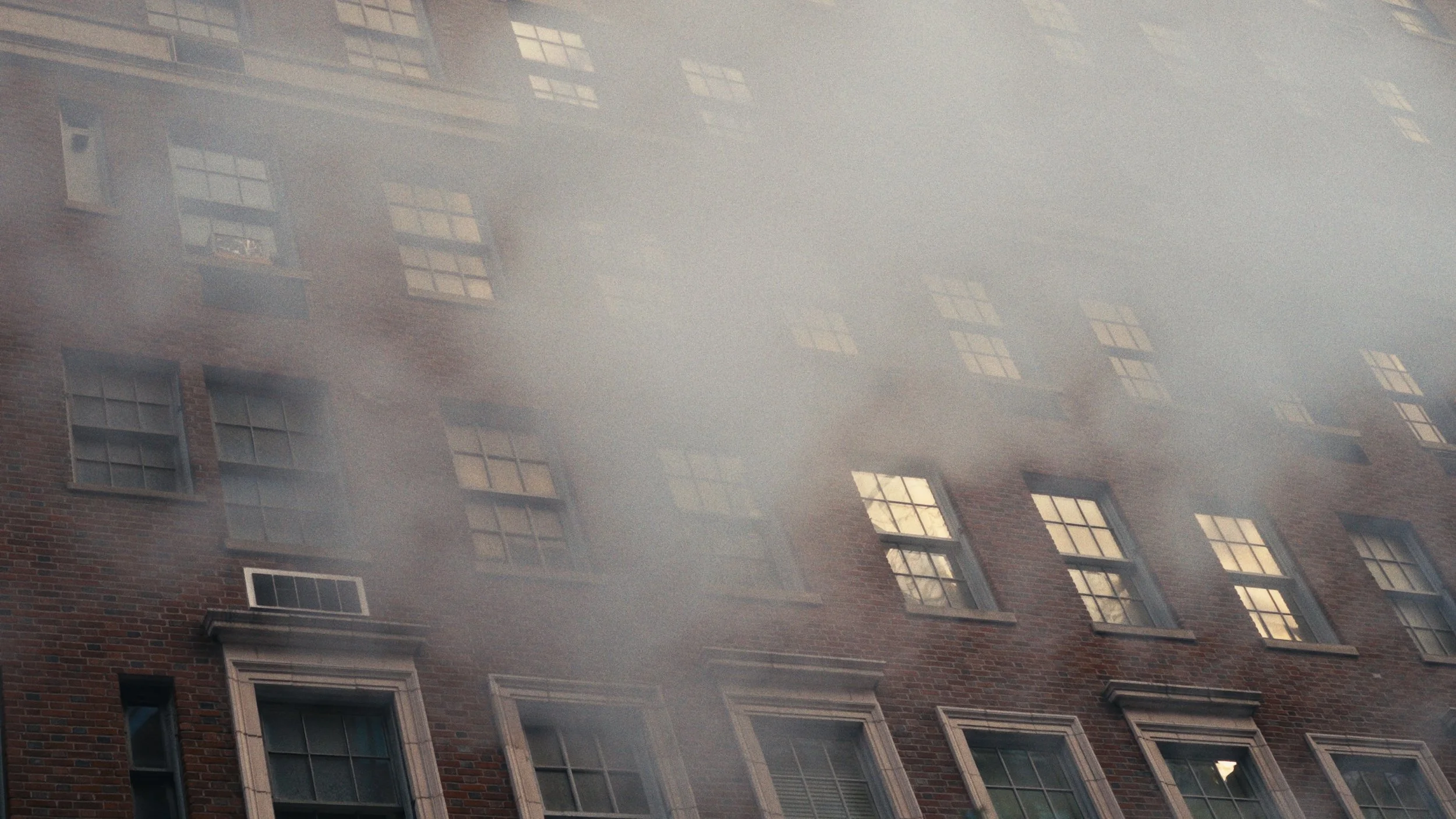 A brick apartment building with multiple windows, some open. The building is reflected in a foggy or steamy surface, creating a hazy overlay.