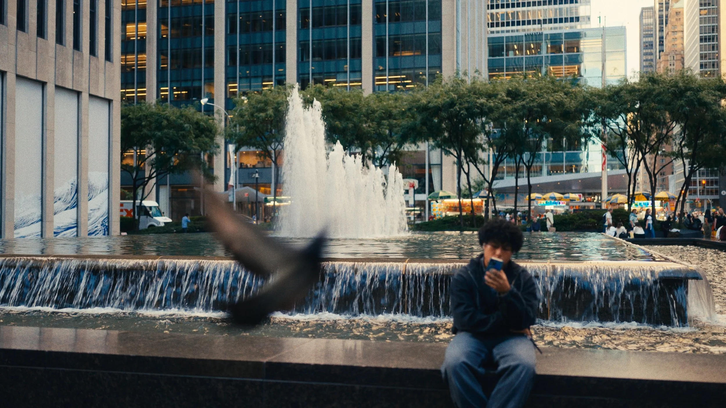A young man sits on the edge of a fountain in an urban plaza, looking at his phone, with a blurred bird flying past in the foreground, surrounded by tall buildings, trees, and city activity.