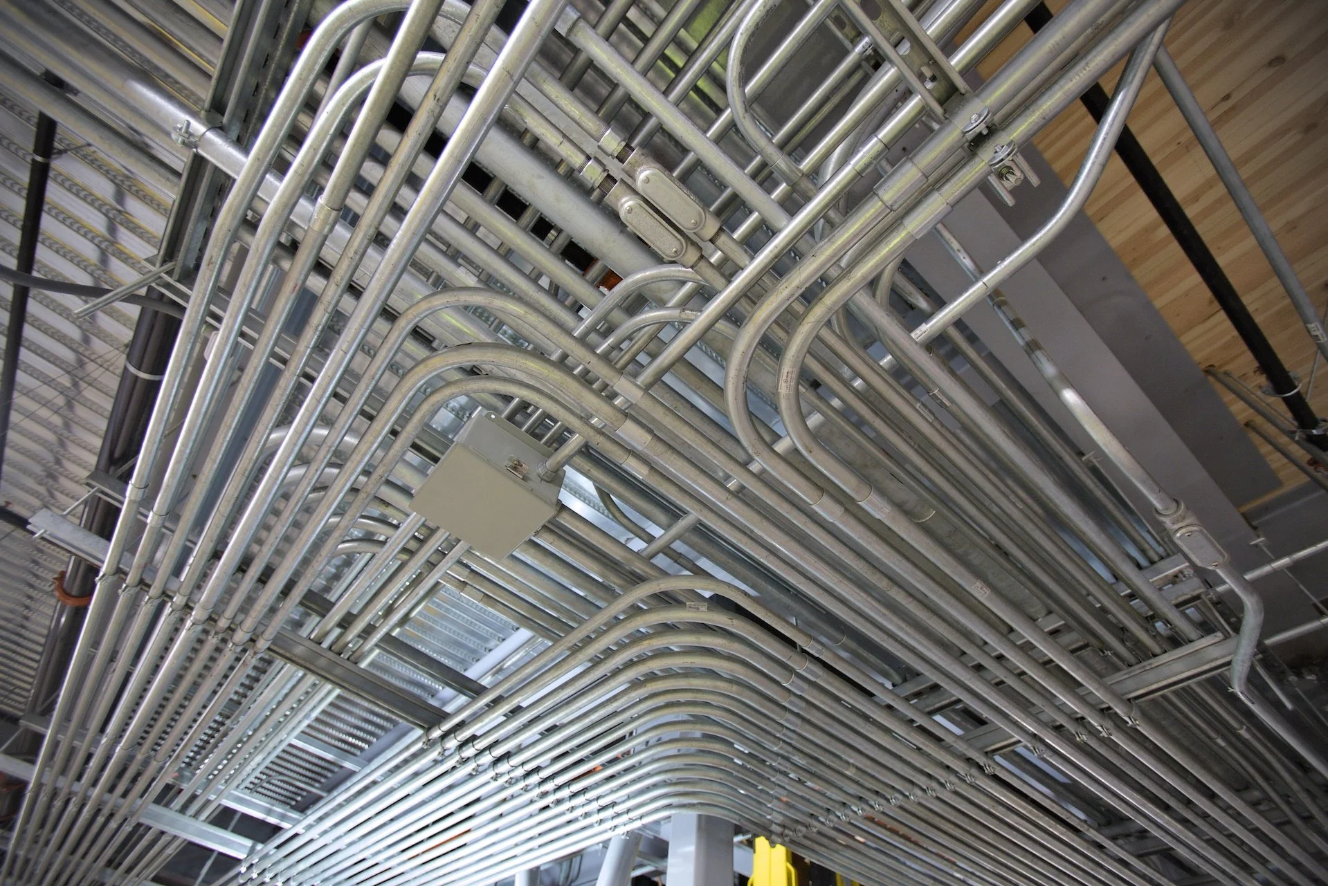 A ceiling with numerous metallic pipes and conduits running in parallel, some curved and some straight, with electrical boxes attached.