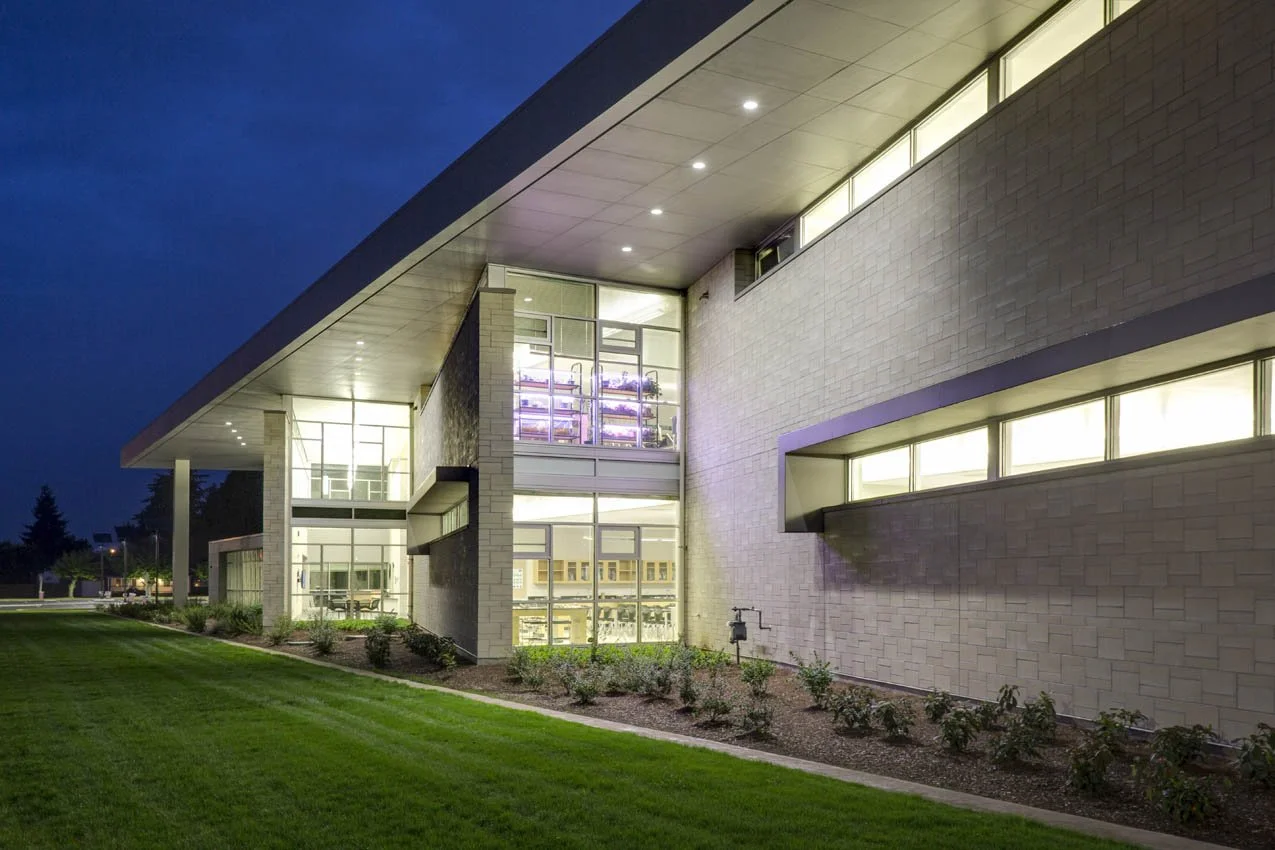 Modern building illuminated at night with large windows, a sloped roof, and landscaped lawn with bushes in the foreground.