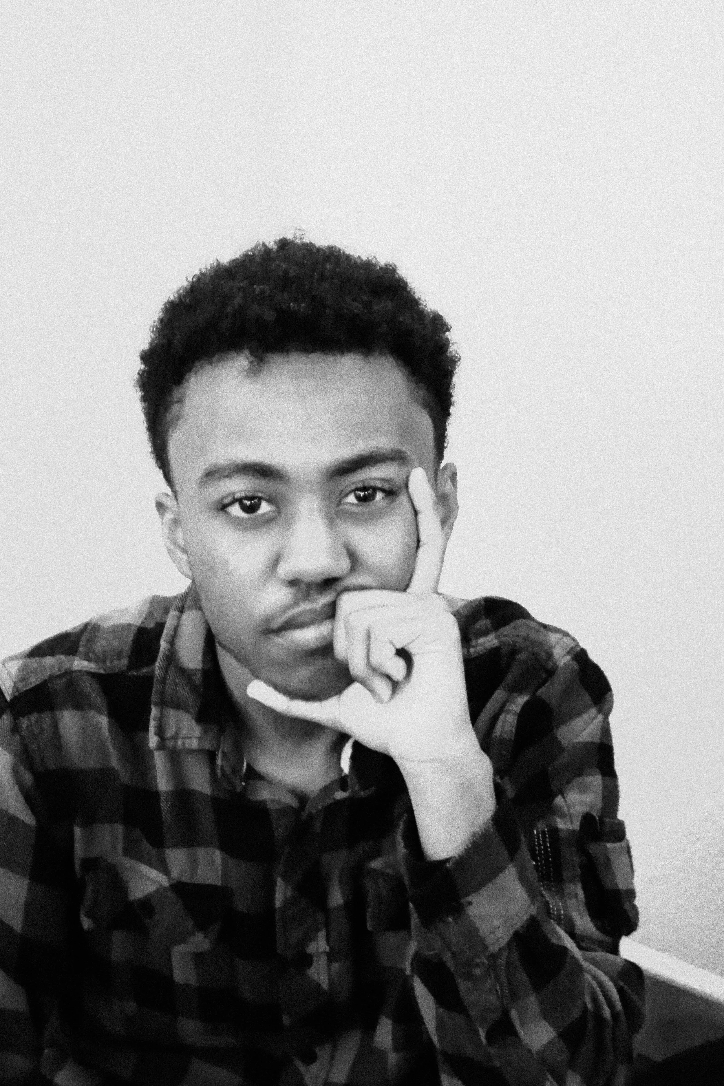 Young man with short, curly hair and a plaid shirt, holding his finger to his temple, looking directly at the camera in black and white.