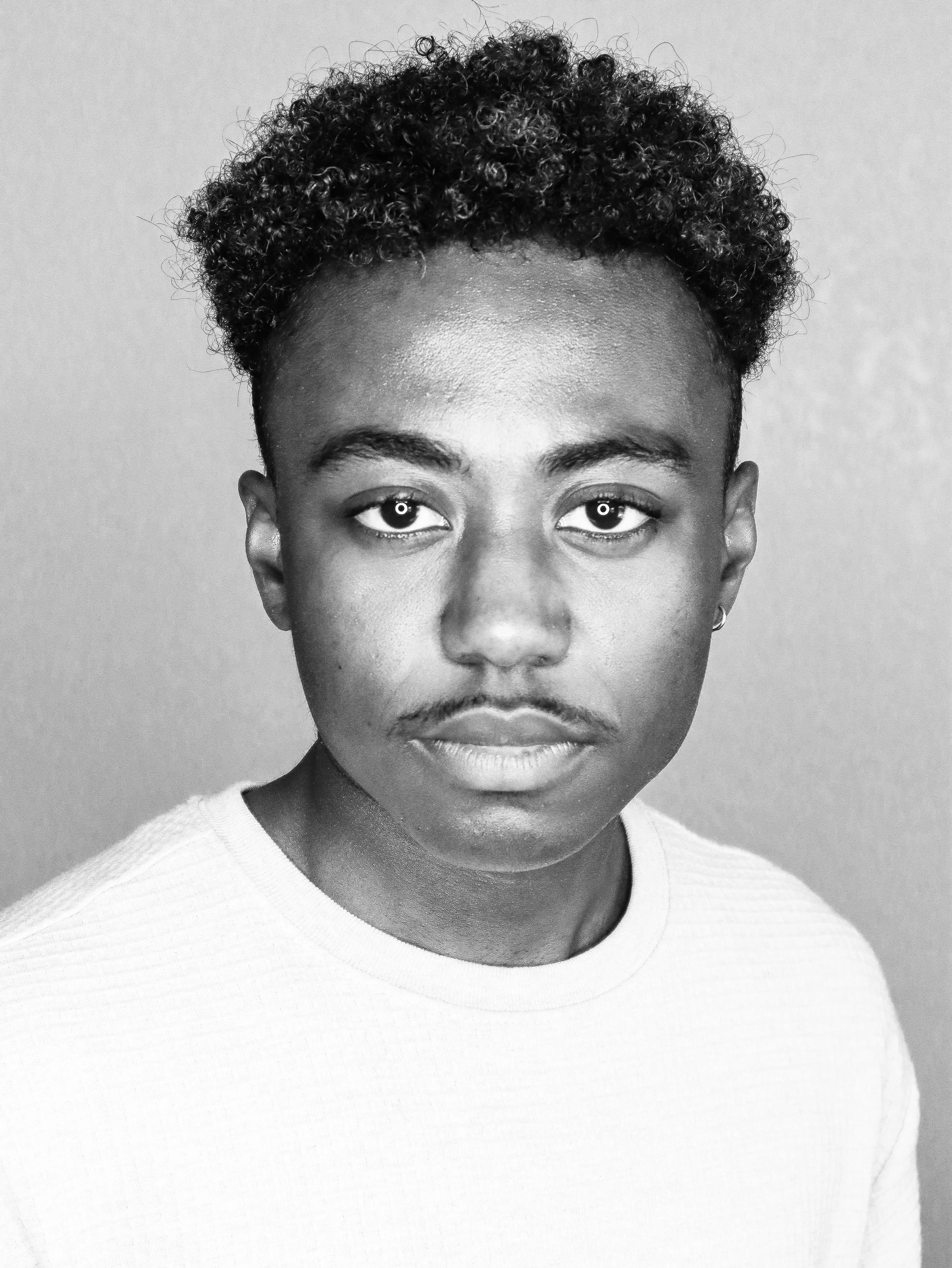 Close-up black and white photograph of a person's face, focusing on the eyes, nose, and mouth. The person has short, curly hair and lightly arched eyebrows, wearing a light-colored shirt.