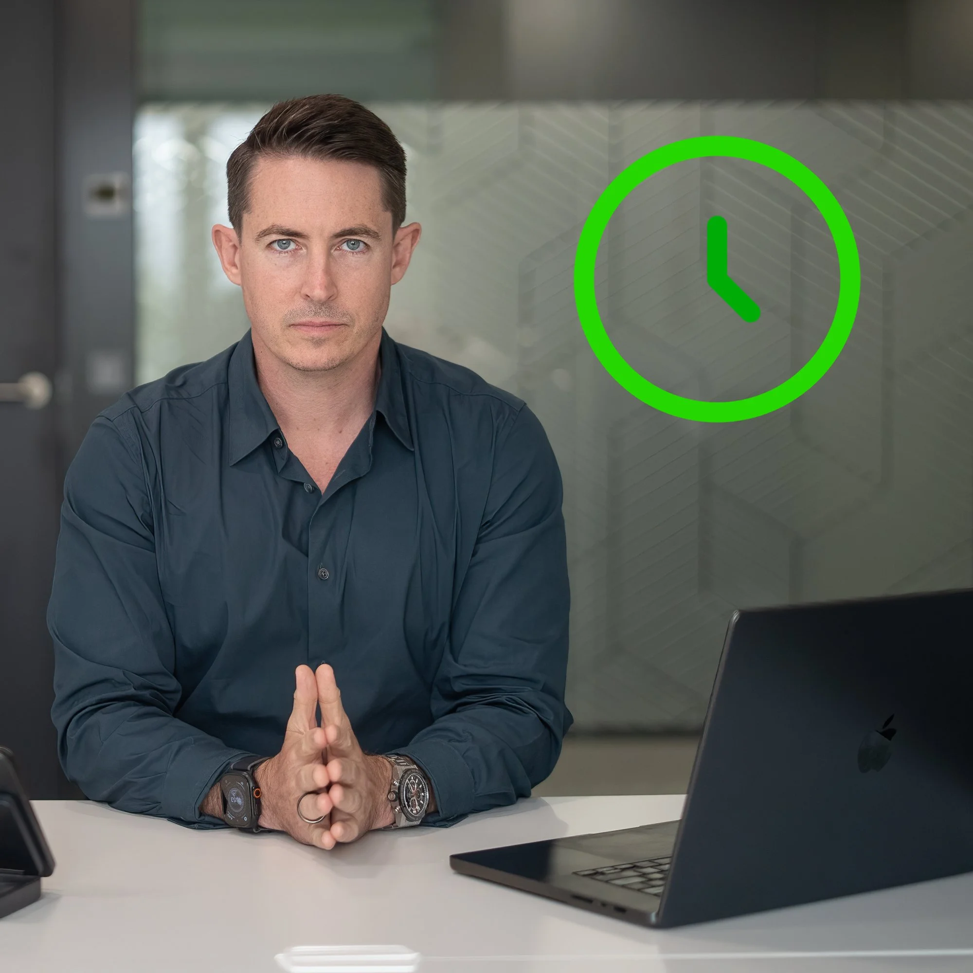 A man sitting at a desk with a laptop, hands clasped, looking at the camera, with a green clock icon on the glass behind him.