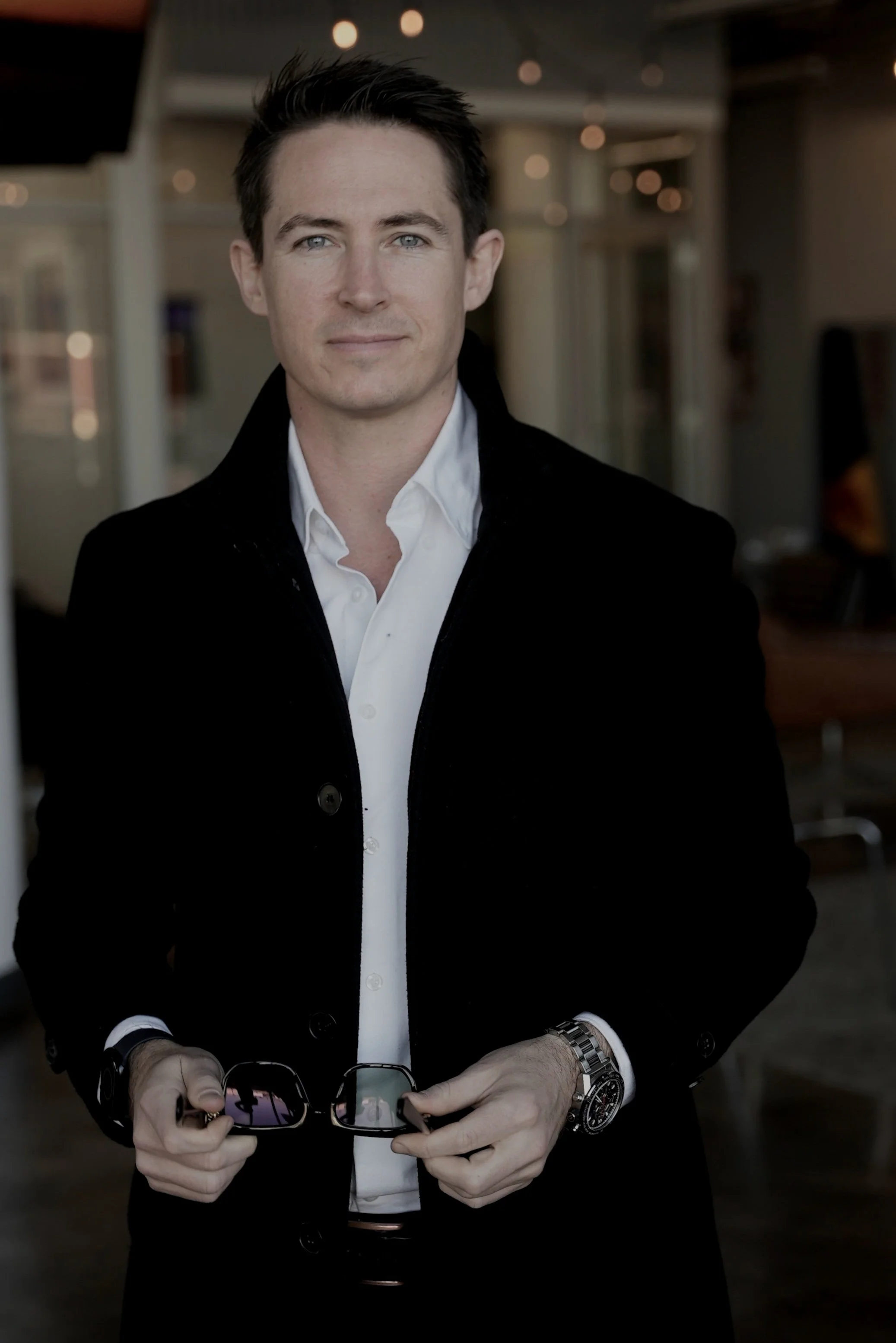 A man with dark hair and blue eyes wearing a white shirt and black coat, holding sunglasses in both hands, standing indoors with blurred background.