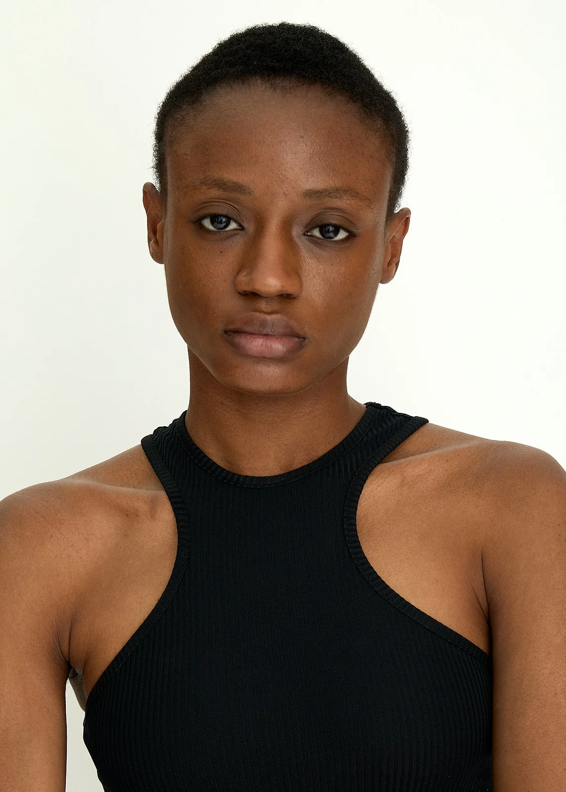 A young Black woman with short hair wearing a black sleeveless top, looking directly at the camera with a neutral expression against a plain white background. Photography by Imani Bahati