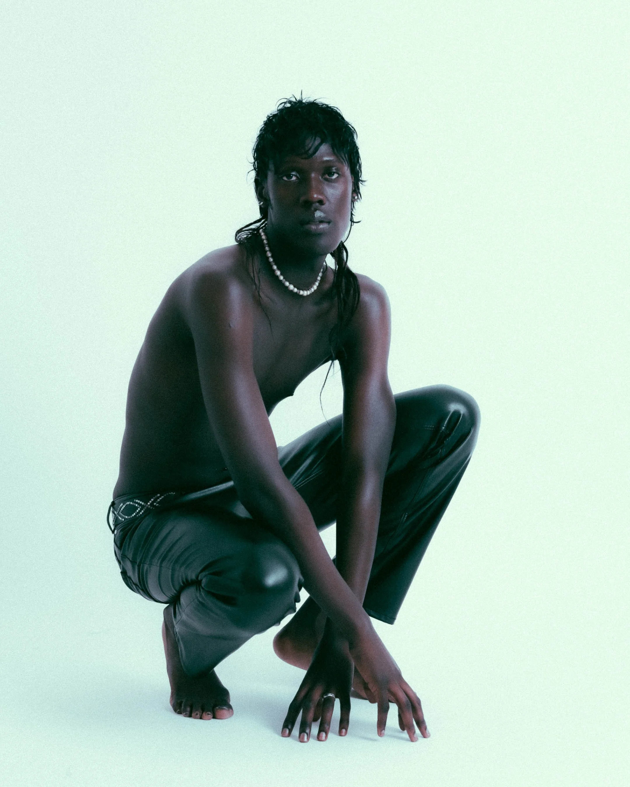 A person with dark skin and wet, black hair crouches on the floor against a plain, light-colored background, wearing leather pants and a pearl necklace. Photography by Imani Bahati