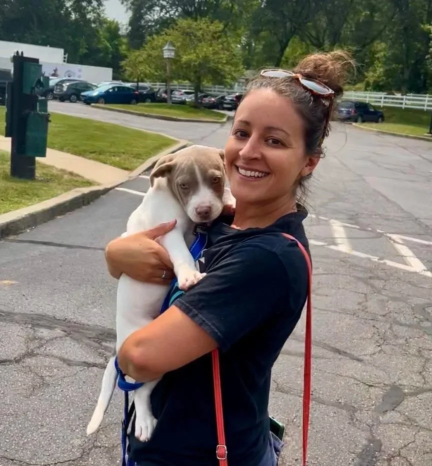 Cori Tufano, owner and head trainer of Lasata Dog Trainer holding a white and brown puppy and walking through a parking lot.