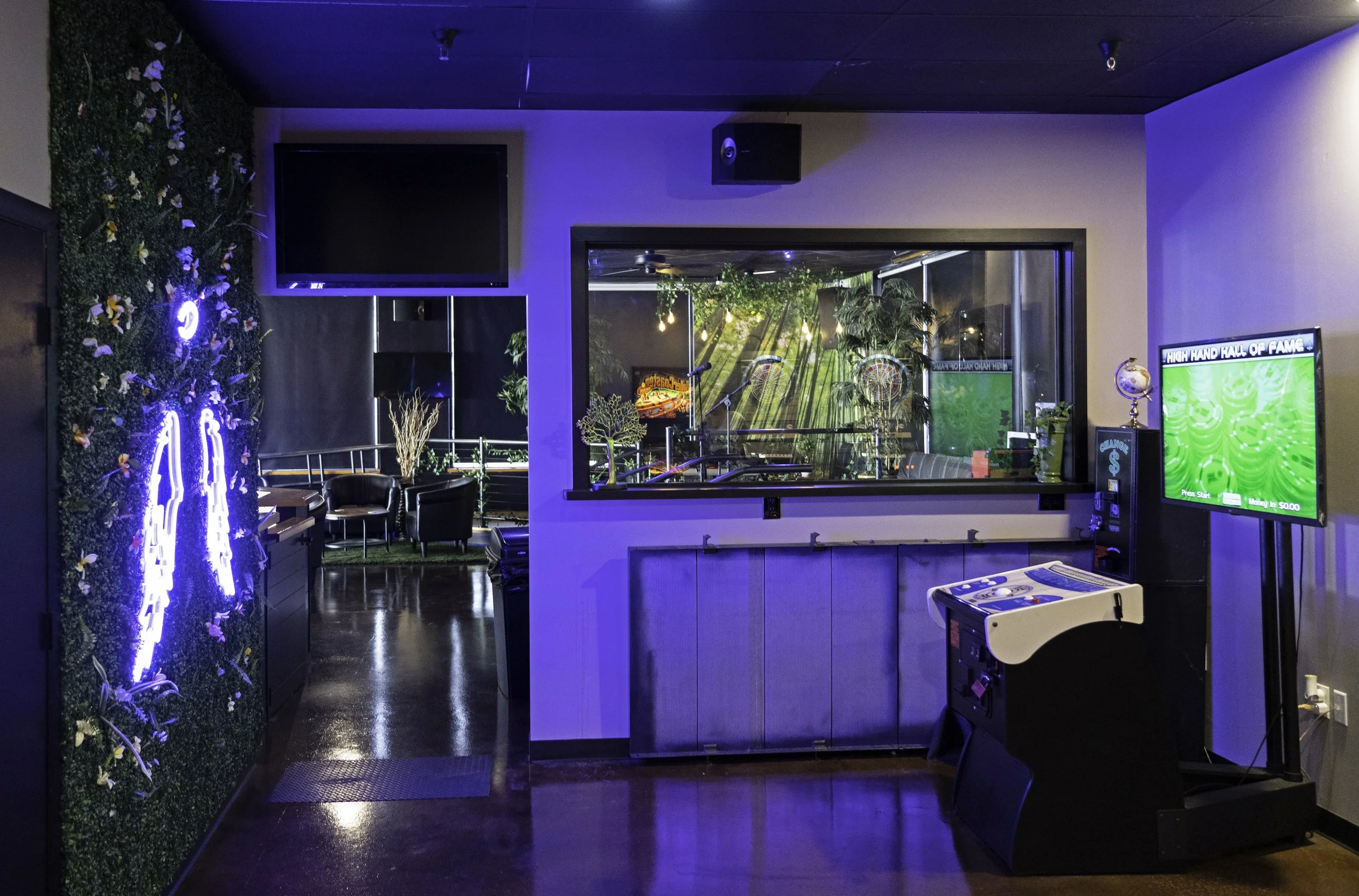 Indoor arcade room with a high score game machine, a small jumbotron screen, a glass window showing a tropical themed outdoor area with jungle decor, gaming souvenirs on top of the high score machine, and a wall with decorative greenery illuminated by blue lighting.