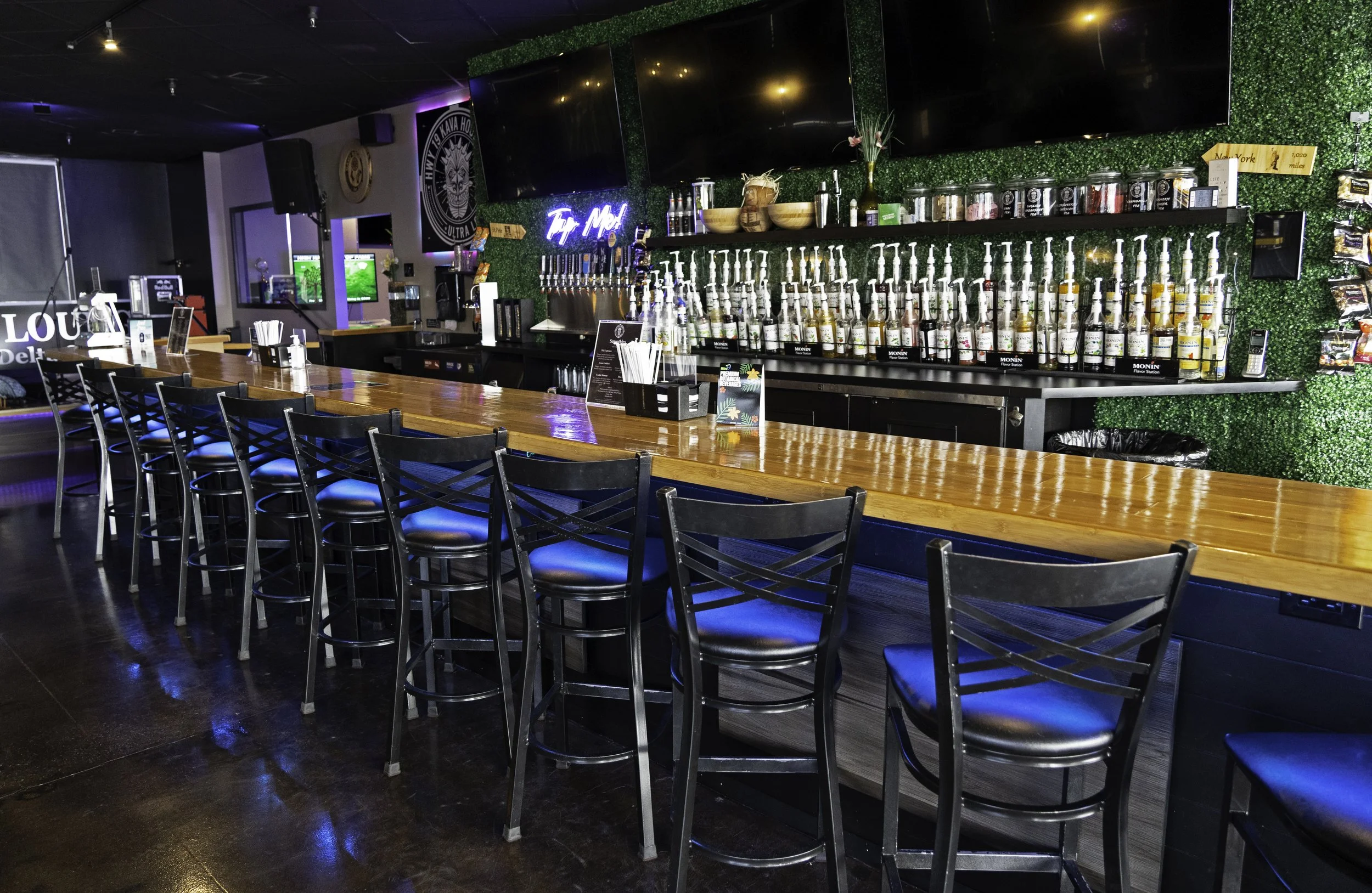 Empty bar with a long wooden counter and black chairs, behind it are liquor bottles on shelves, with neon signs and TVs on the walls of a dimly lit bar or nightclub.