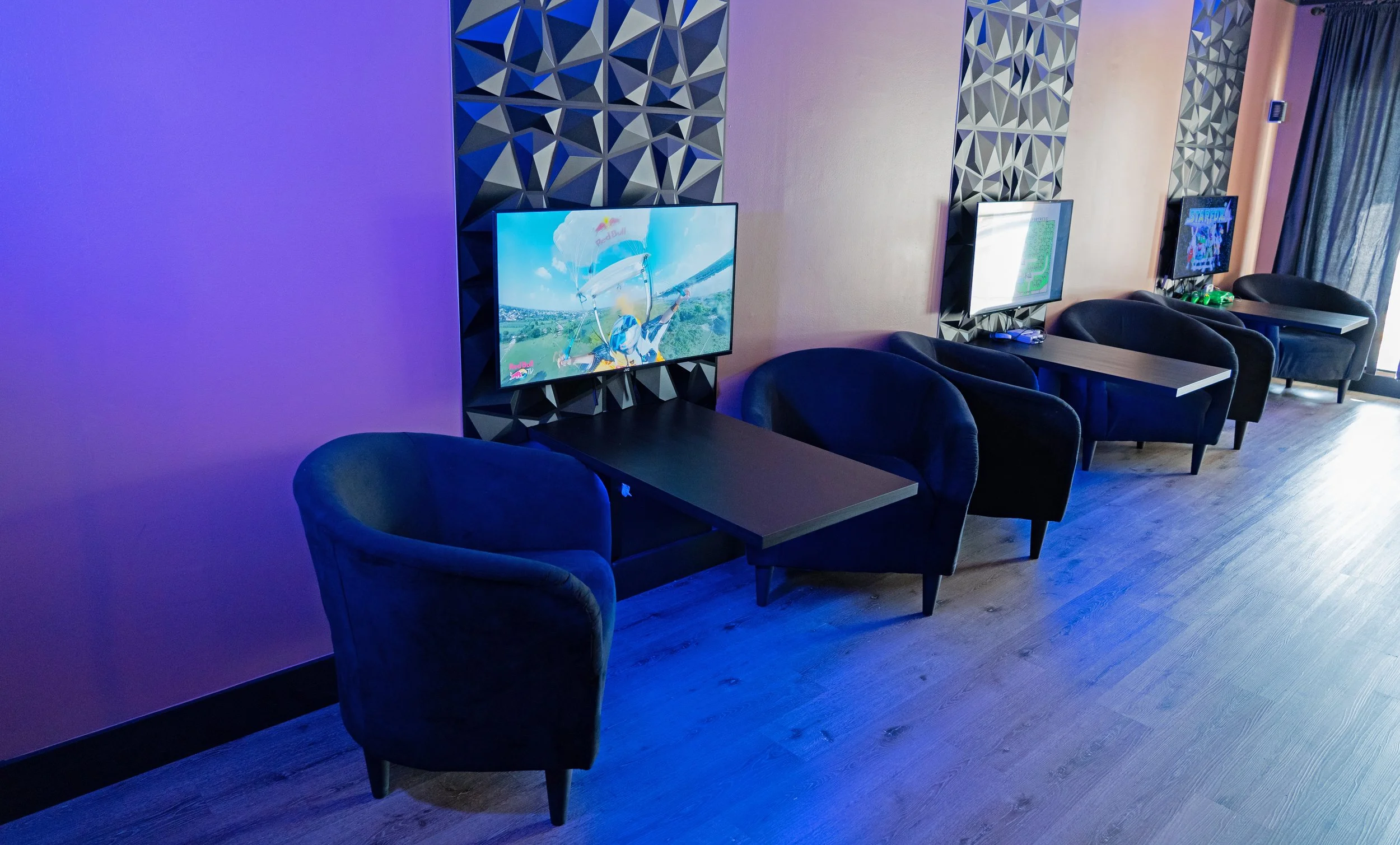 A row of gaming chairs with screens mounted behind them, in a lounge area with blue lighting and wooden flooring.