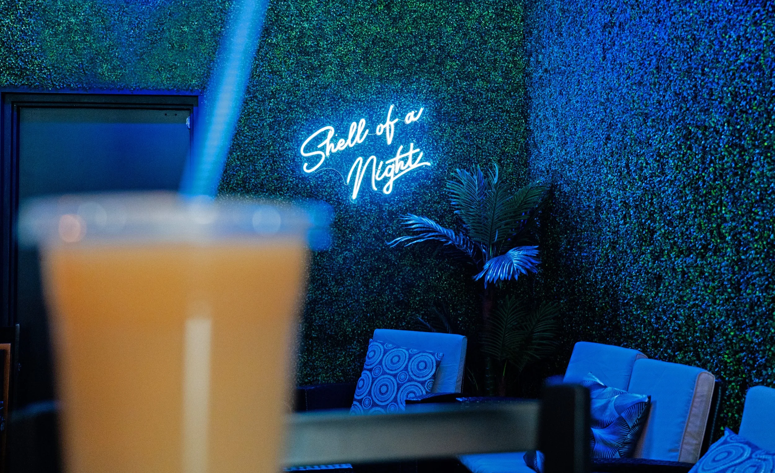 Neon sign reading 'Shell of a Night' on a green textured wall, with blue lighting. Part of a glass with a straw in the foreground and seating with patterned pillows and a potted plant in the background.