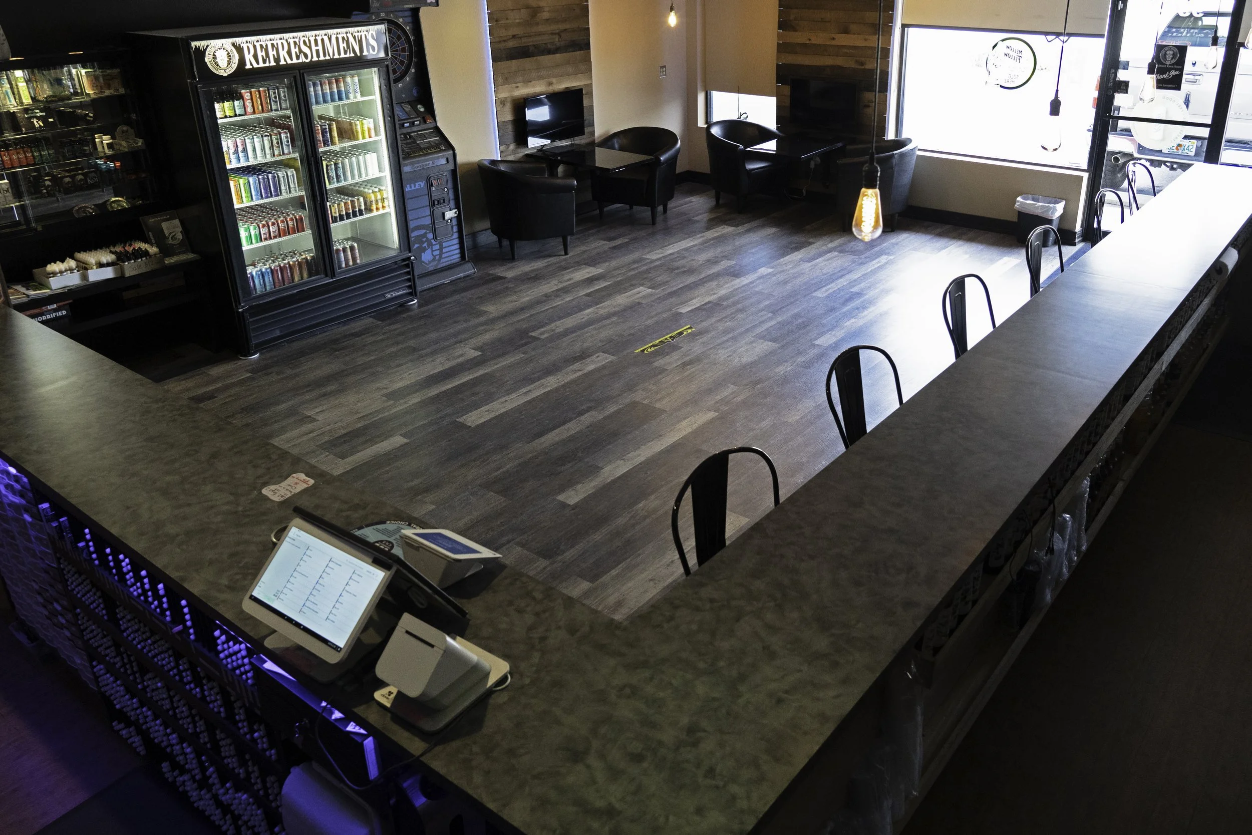 Empty bar counter with barstools, a cash register, and a digital order screen. Behind the counter are a drink fridge, seating area with black chairs and tables, a window with sunlight, and a door leading outside.