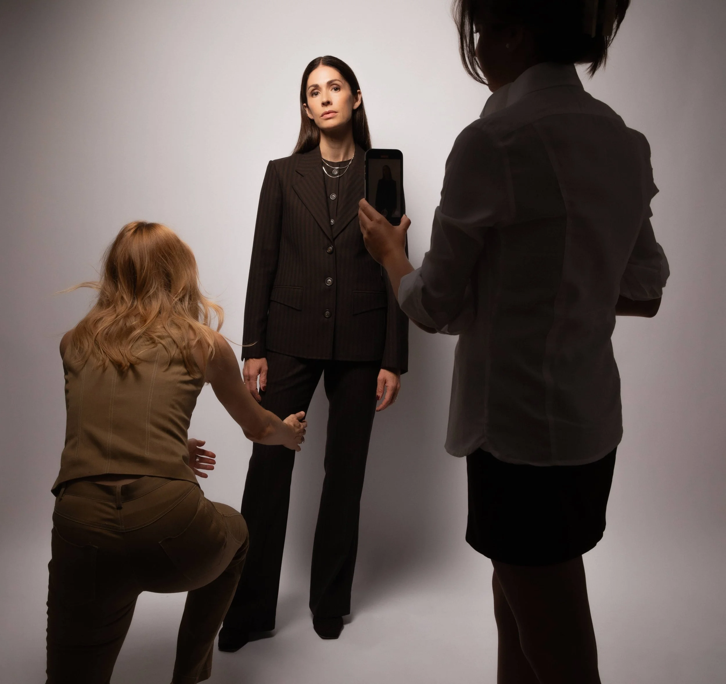 Danu Hassik Kennedy of Known Work wearing a black and brown stripe three piece suit standing against a plain background while two women, one with red hair kneeling and one with dark hair standing, take photos of her and fit her in her suit.