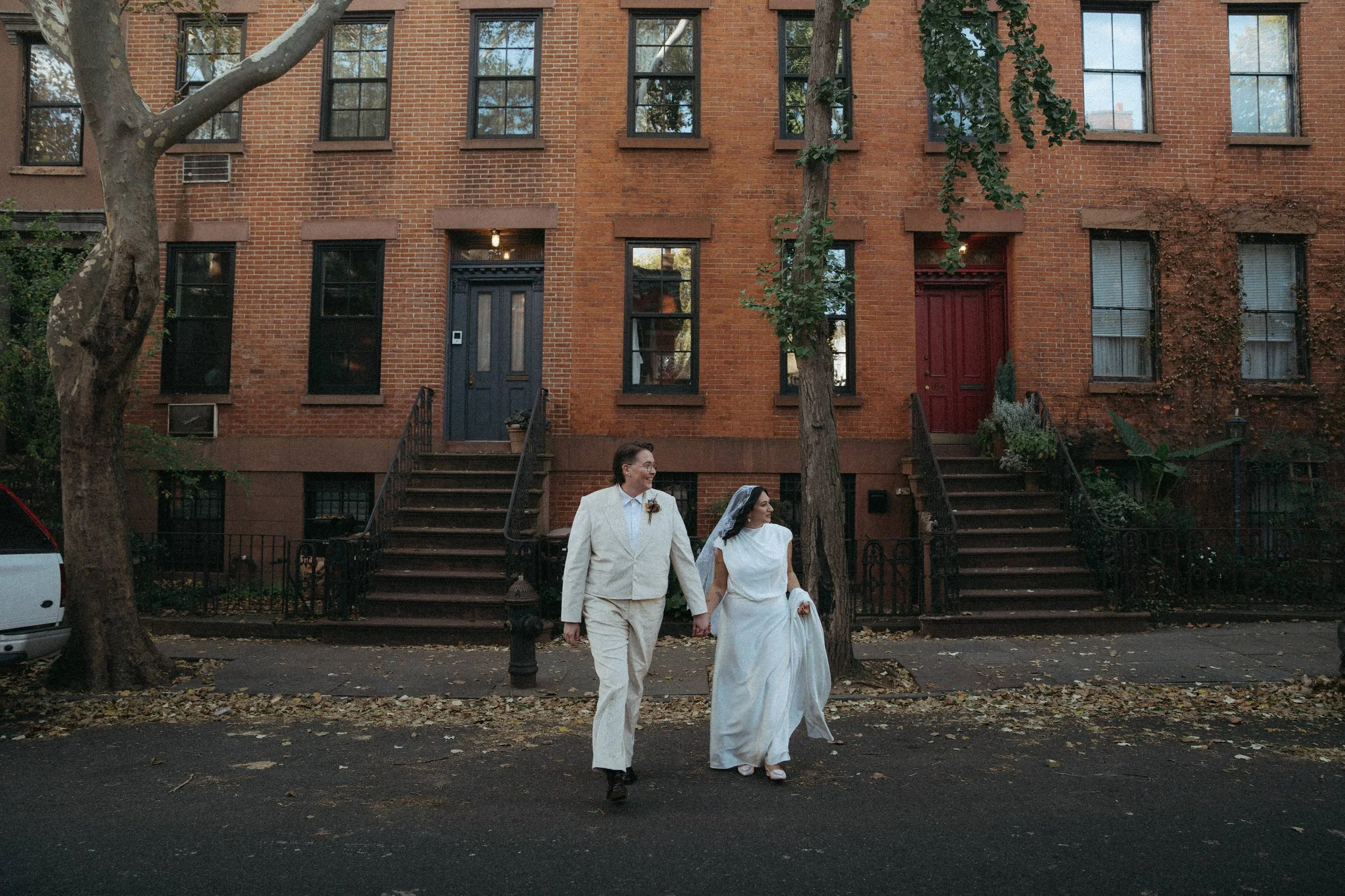 A couple dressed in wedding attire walking on a city street in front of brown brick apartment buildings with stairs leading up to their entrances. The woman is wearing a white wedding dress and nonbinary person in a cream-colored velvet moire suit