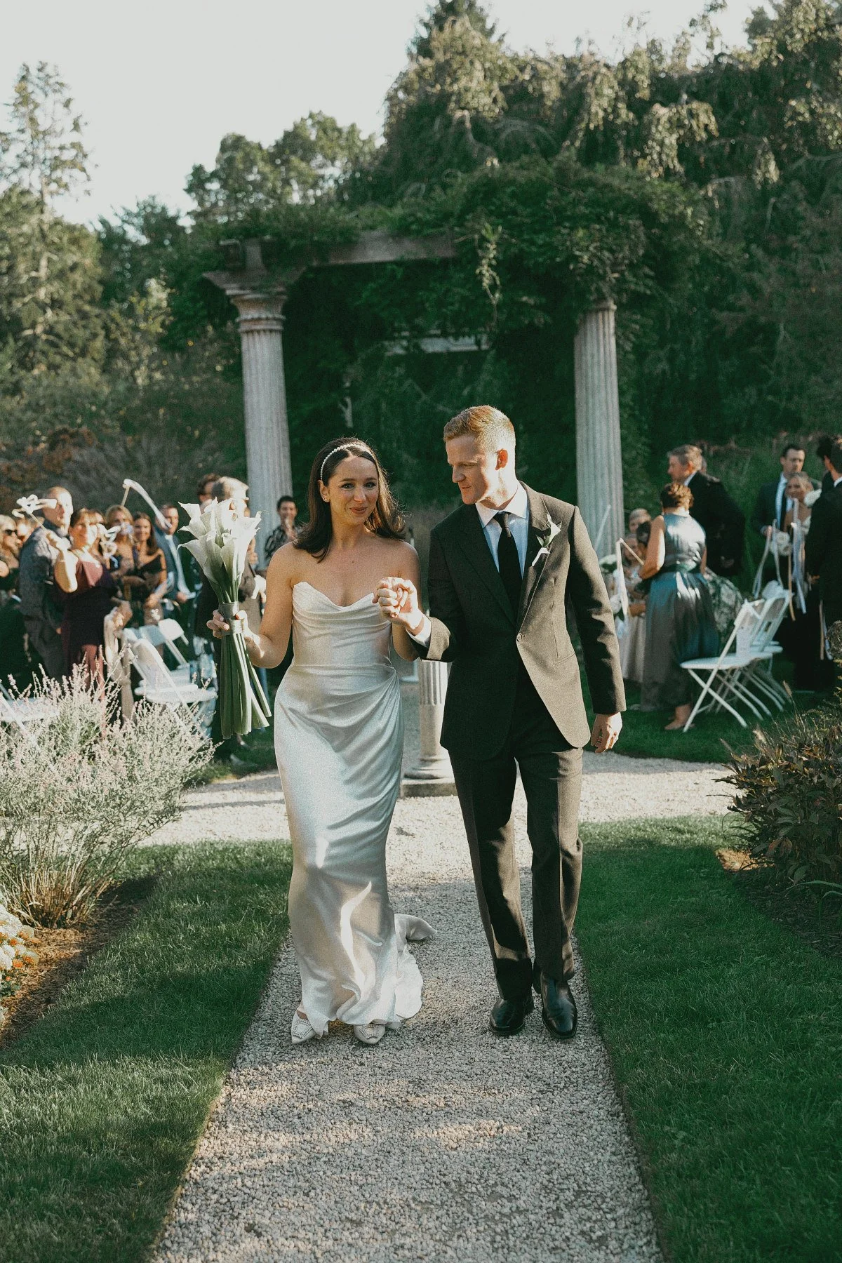 A bride and groom walking hand in hand down an outdoor aisle during a summer wedding ceremony, the groom is wearing a green custom tailored watson ellis wedding suit.