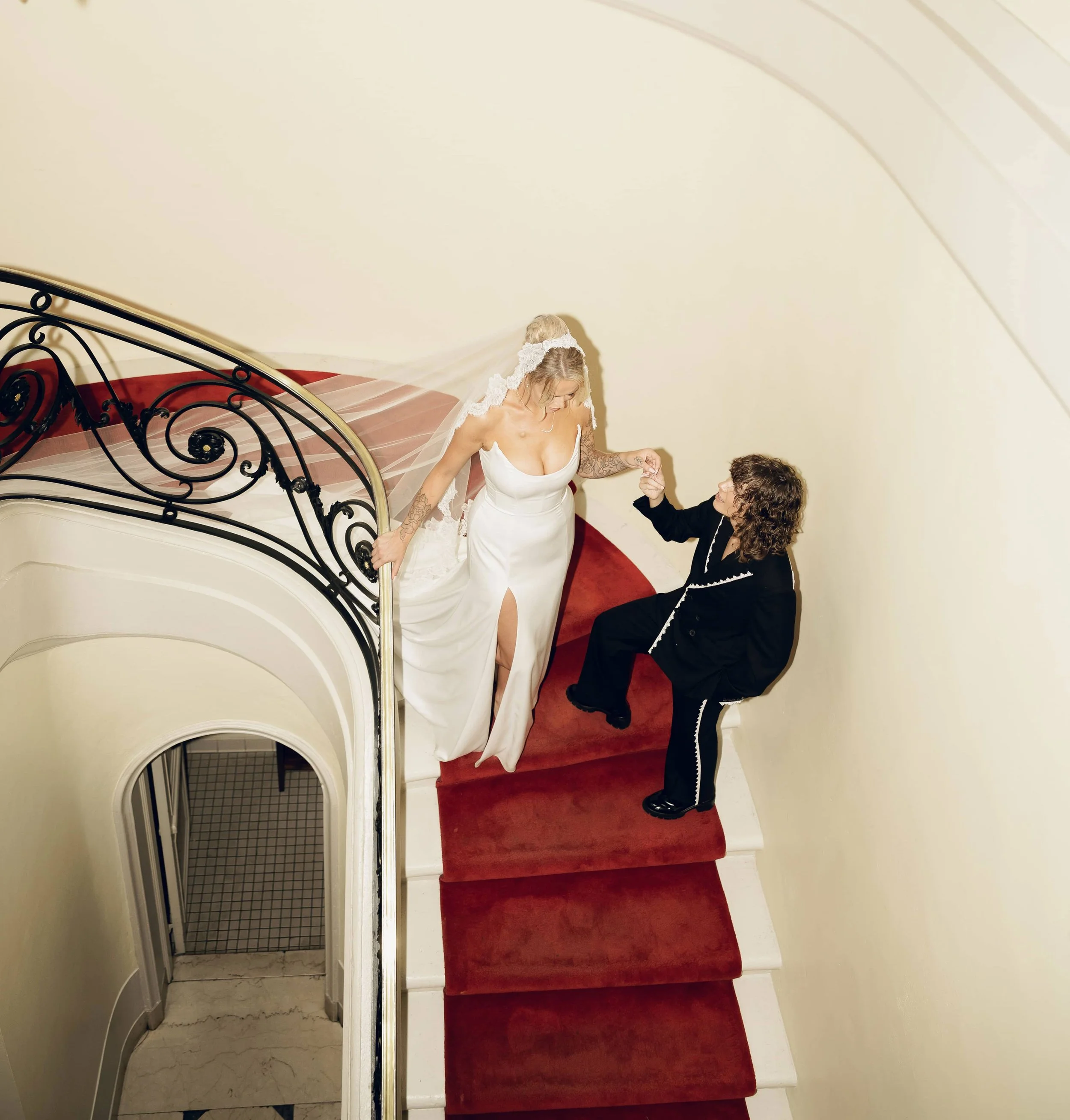 A lesbian wedding picture of a bride in a white wedding dress holding hands with a bride in a black suit with white lace detail on a red carpeted staircase.