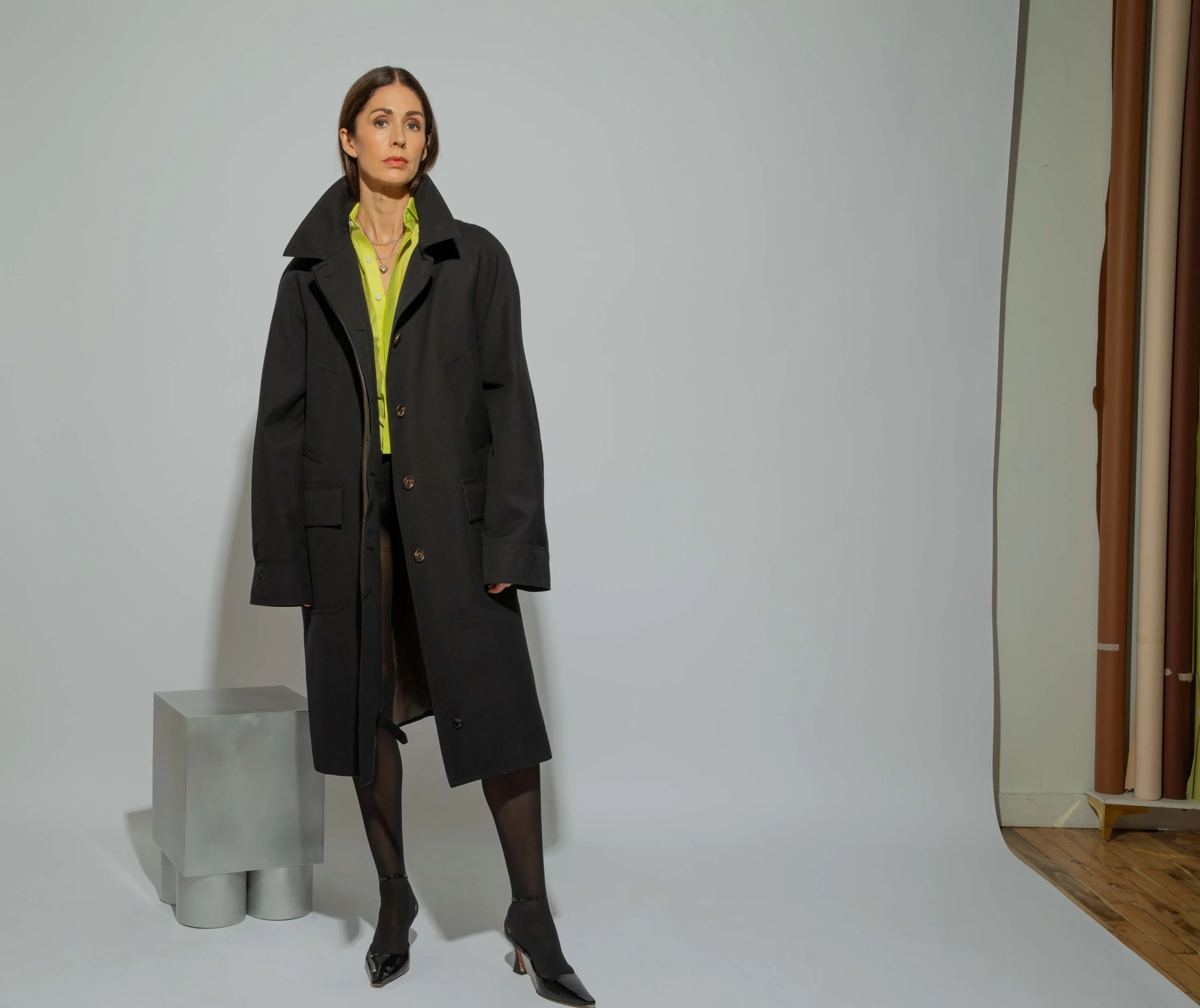 A woman standing against a plain gray background, wearing a long black coat, green shirt, black tights, and black high heels, with a neutral expression.