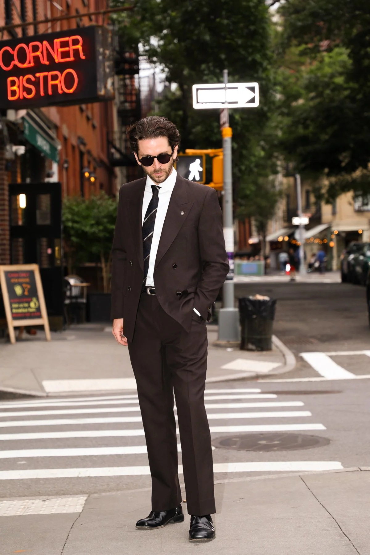 A man dressed in a chocolate brown suit, white shirt, striped navy tie, and sunglasses standing on a crosswalk in the west village of NYC with the bar's Corner Bistro sign behind him.