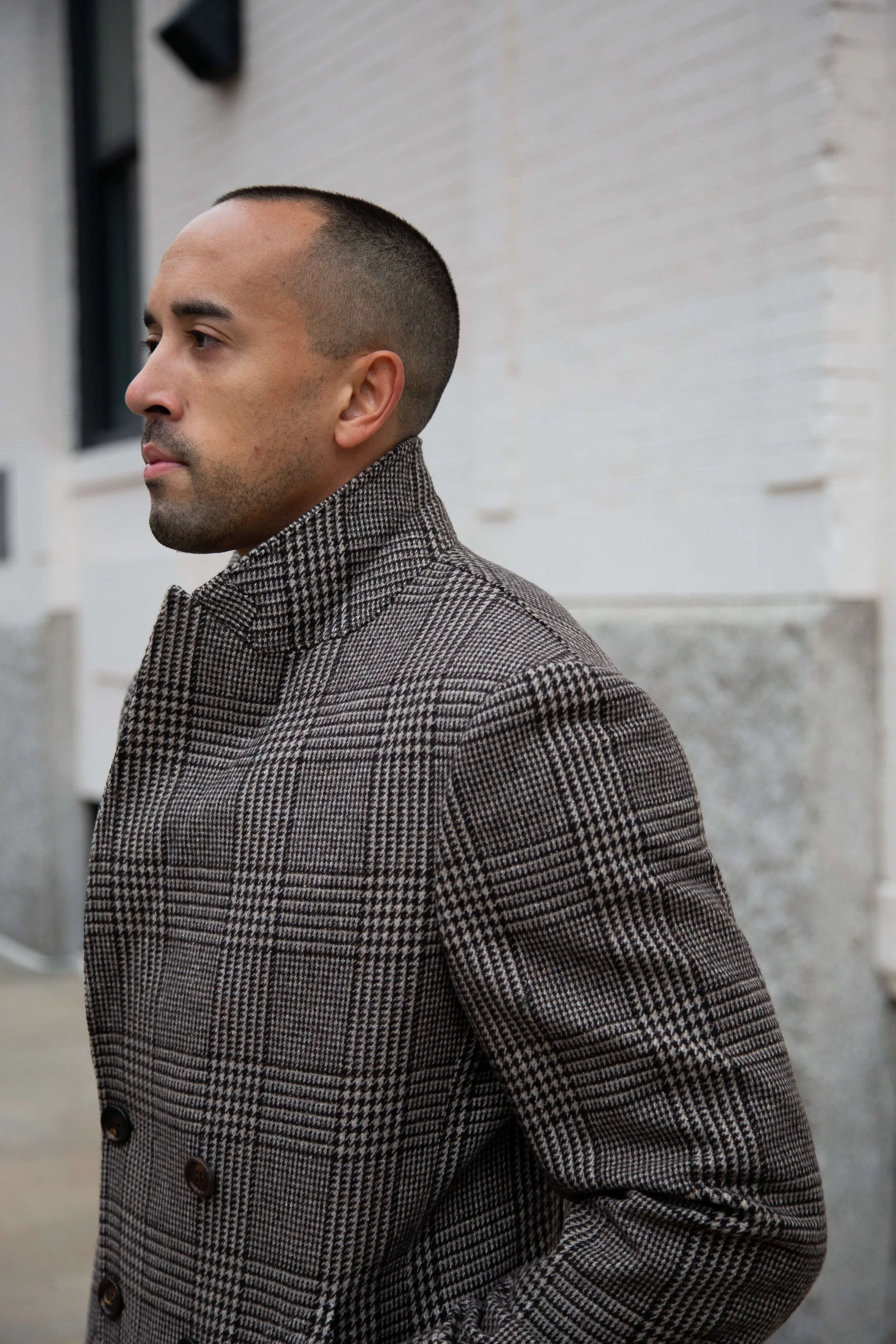A man with a buzz cut hairstyle wearing a brown plaid coat stands outside against a white brick wall.