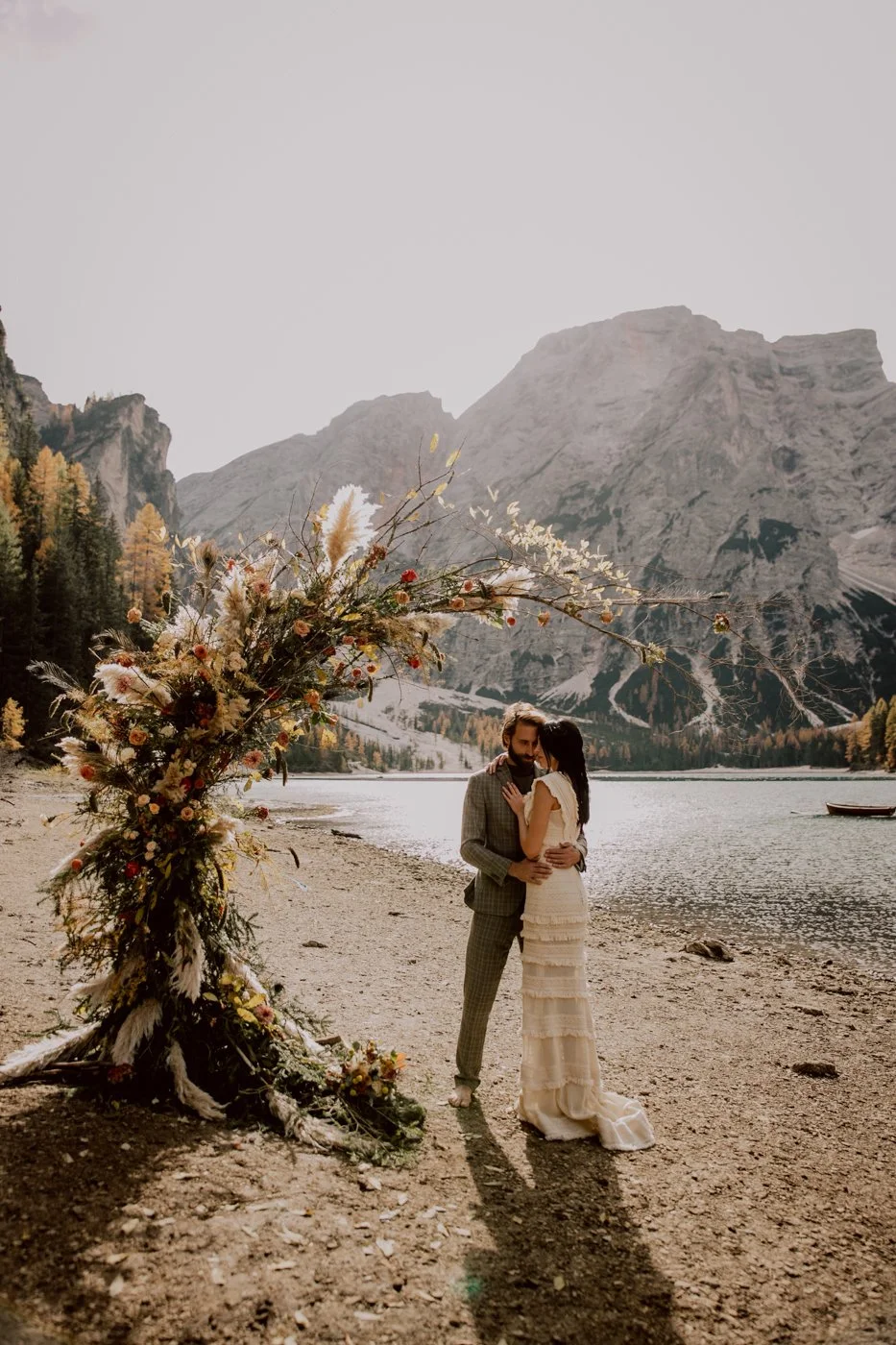 A couple in wedding attire standing on a lakeshore, embracing, with a mountain backdrop and a floral arrangement nearby.