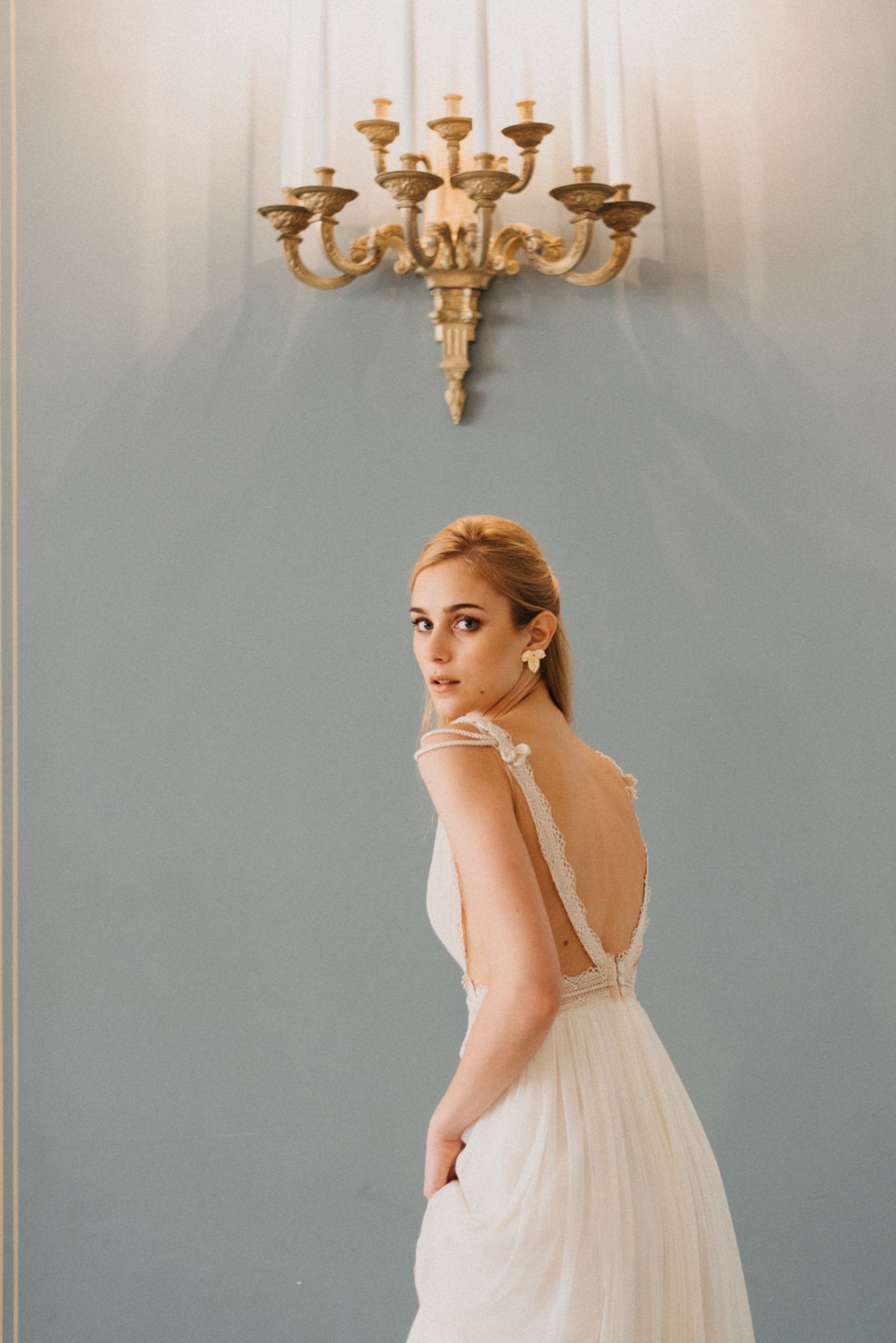 Elegant woman in a white dress with lace details, standing in a softly lit room under a vintage chandelier, against a blue wall.
