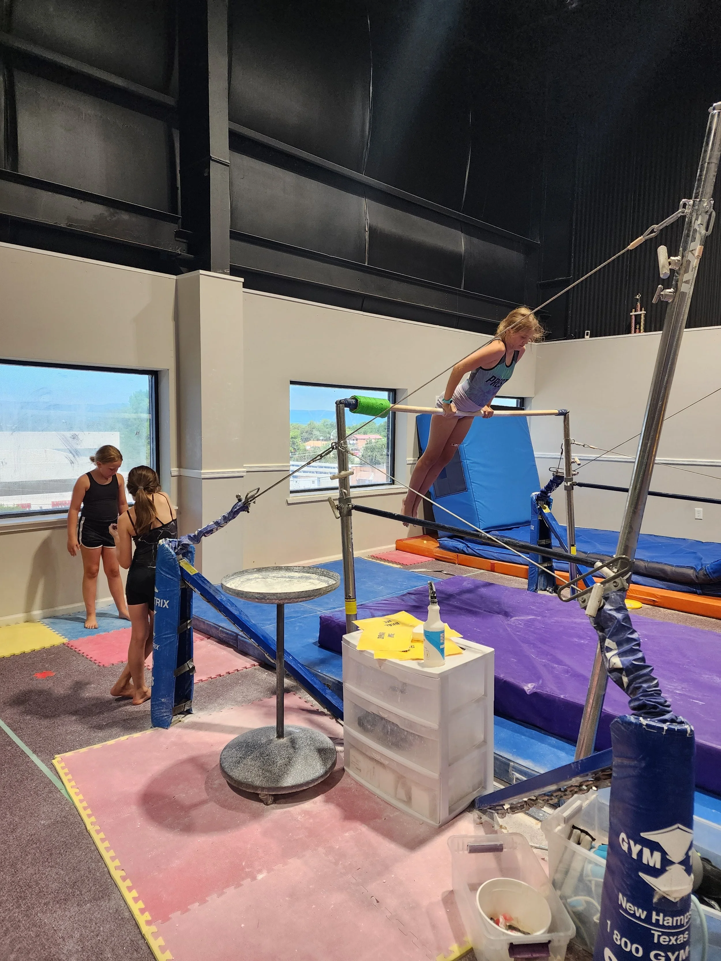 A female gymnast practicing on the uneven bars in a gymnasium, with coaches and other gymnasts nearby.