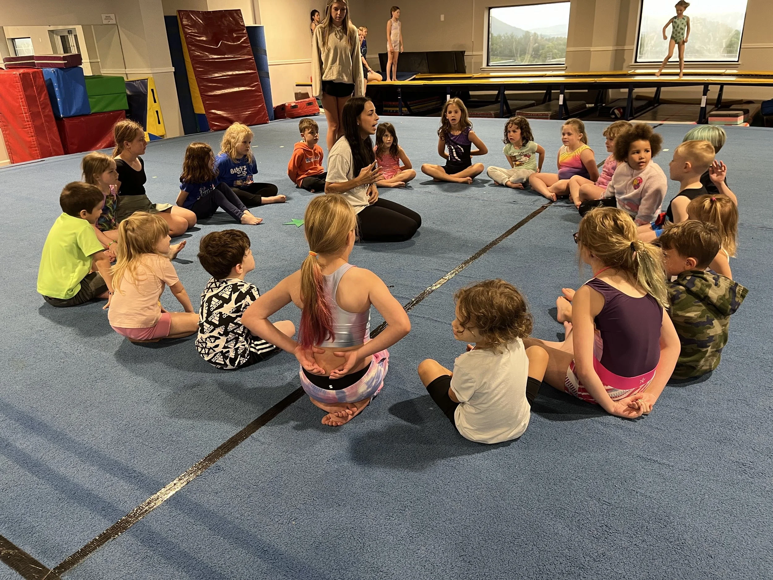 Group of young children sitting in a circle on a blue gymnastics mat listening to an instructor during a class. Indoor gym with large windows, climbing equipment, and a few adults in the background.