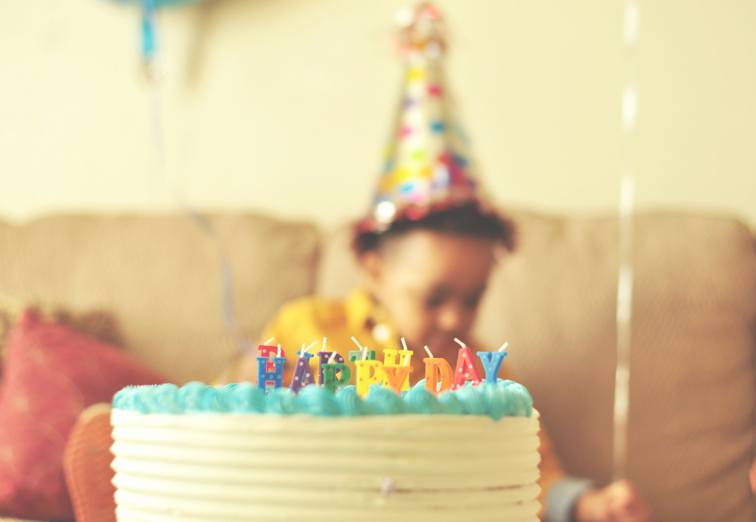 Birthday cake with blue frosting and colorful candles spelling 'HAPPY BIRTHDAY' in front of a person wearing a party hat, sitting on a sofa.