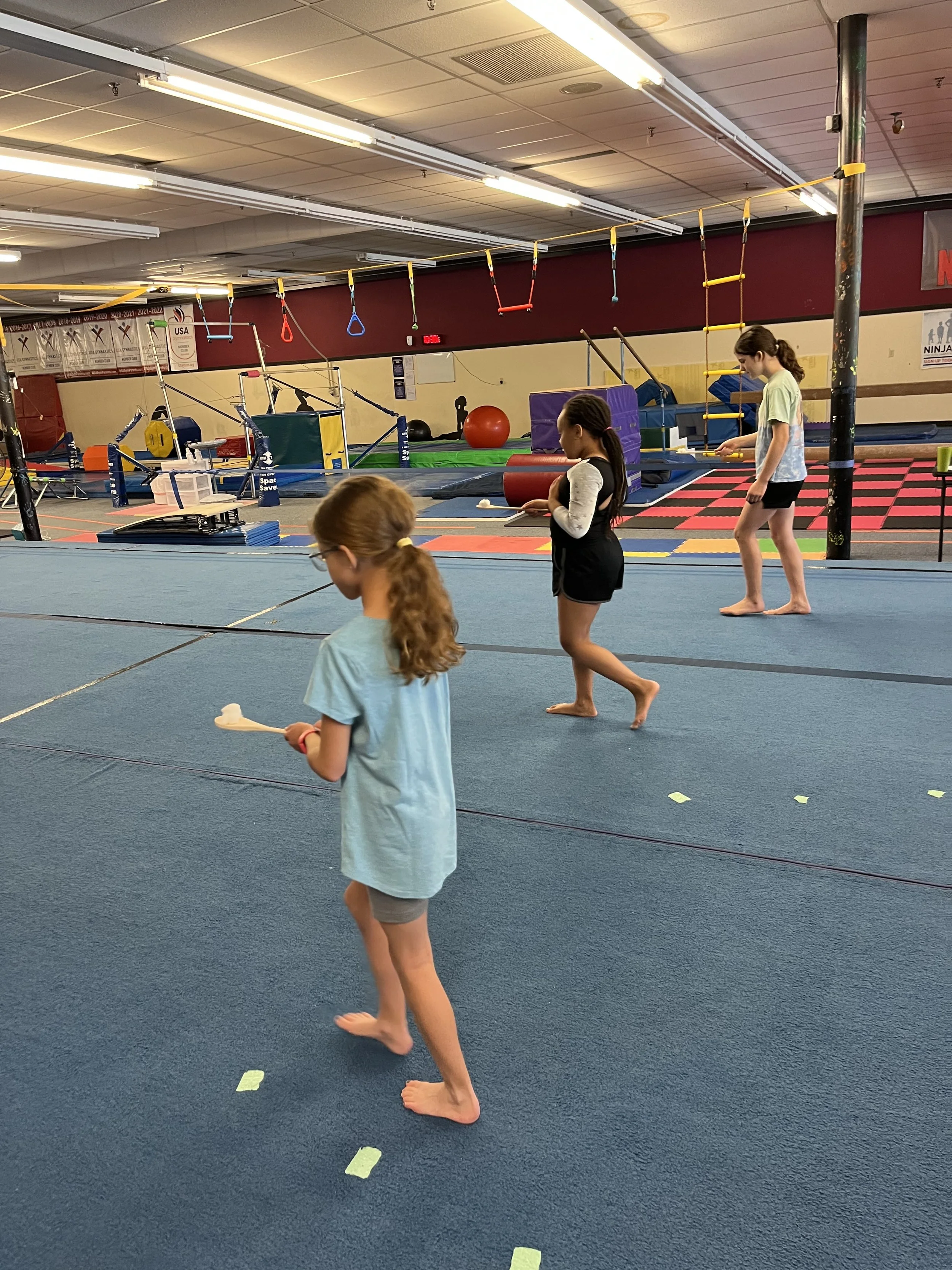 Three children practicing gymnastics in an indoor gym, with one girl holding a small bat or racket in the foreground and two other girls standing at a distance, all barefoot on a blue mat. The background shows gymnastics equipment and hanging rings.