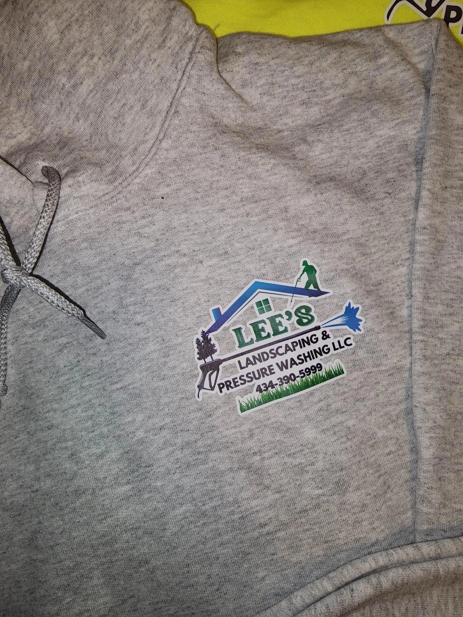Gray hoodie with a logo for Lee's Landscaping & Pressure Washing LLC, featuring a house with a roof, a person pressure washing, and a grass patch with a phone number.
