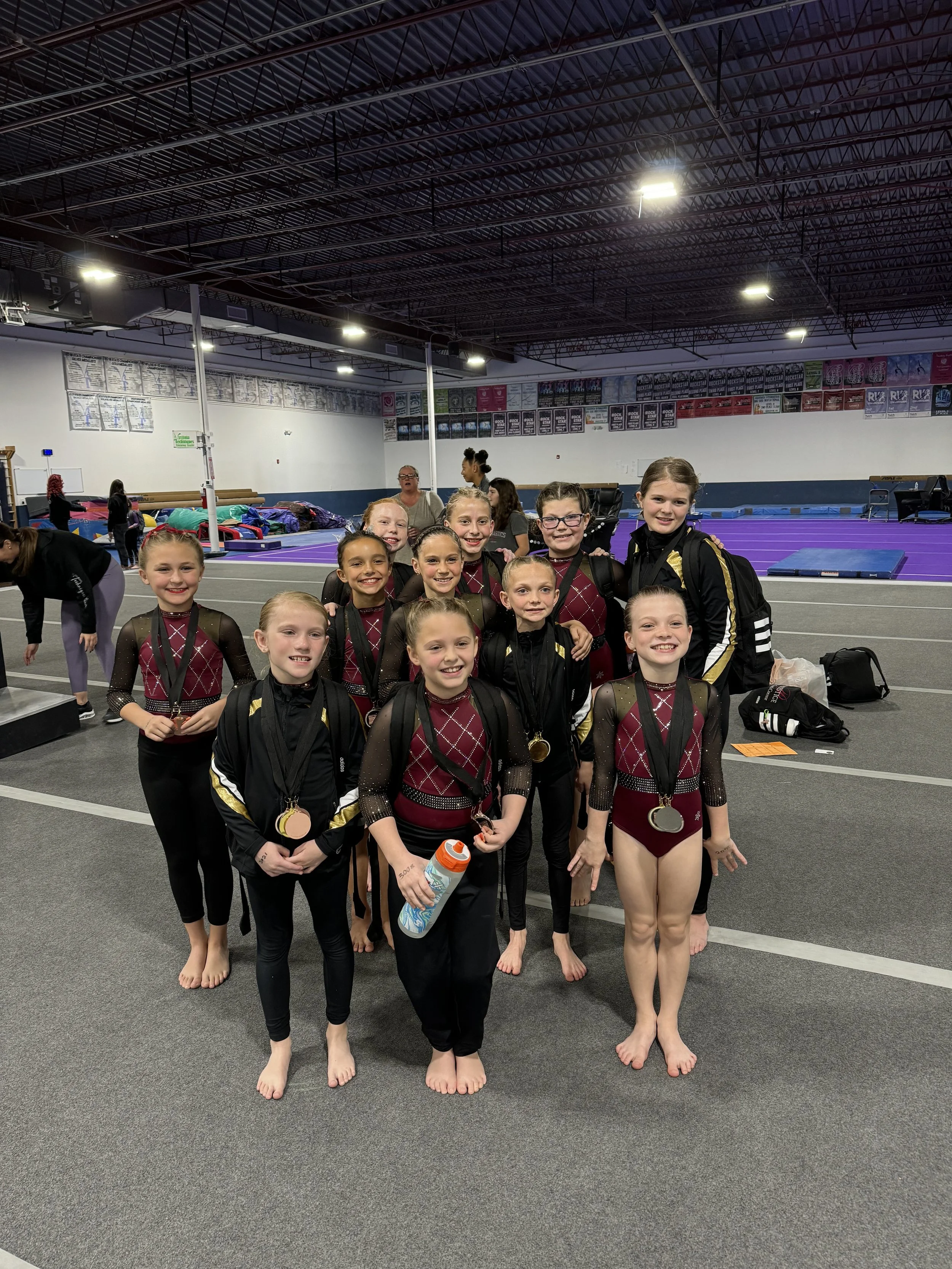 A group of young female gymnasts with medals around their necks standing in a gymnasium, smiling after a competition.