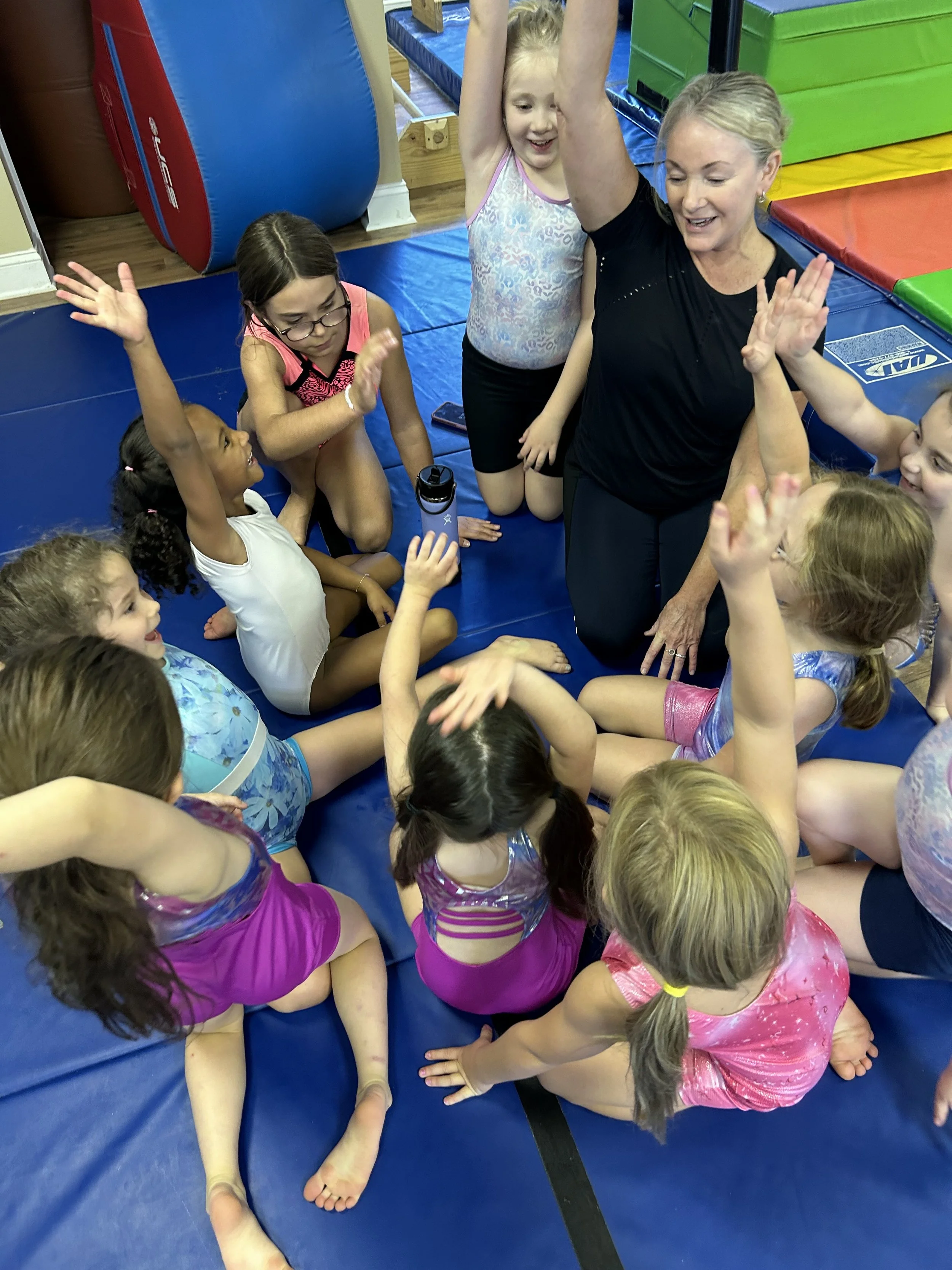 Group of young girls and a woman engaging in a gymnastics or dance activity on a blue mat, with colorful gym equipment in the background.