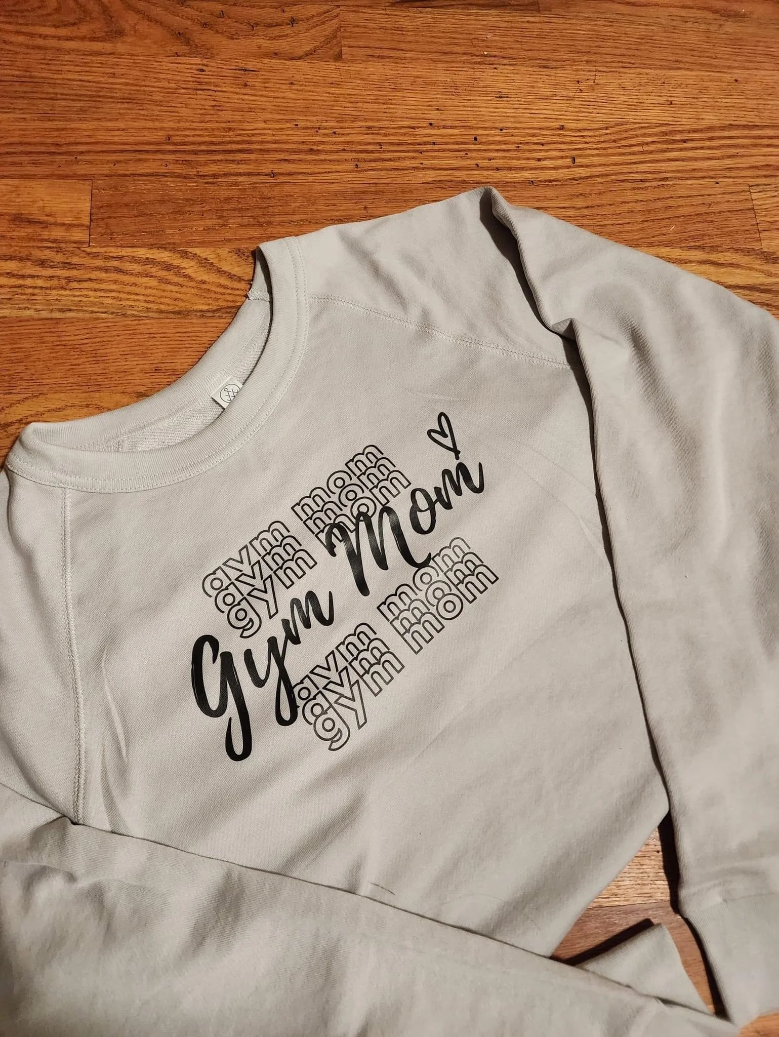 A light-colored sweatshirt with the word "gym" repeatedly printed in small font and "Mom" written in large cursive with a heart, lying on a wooden floor.
