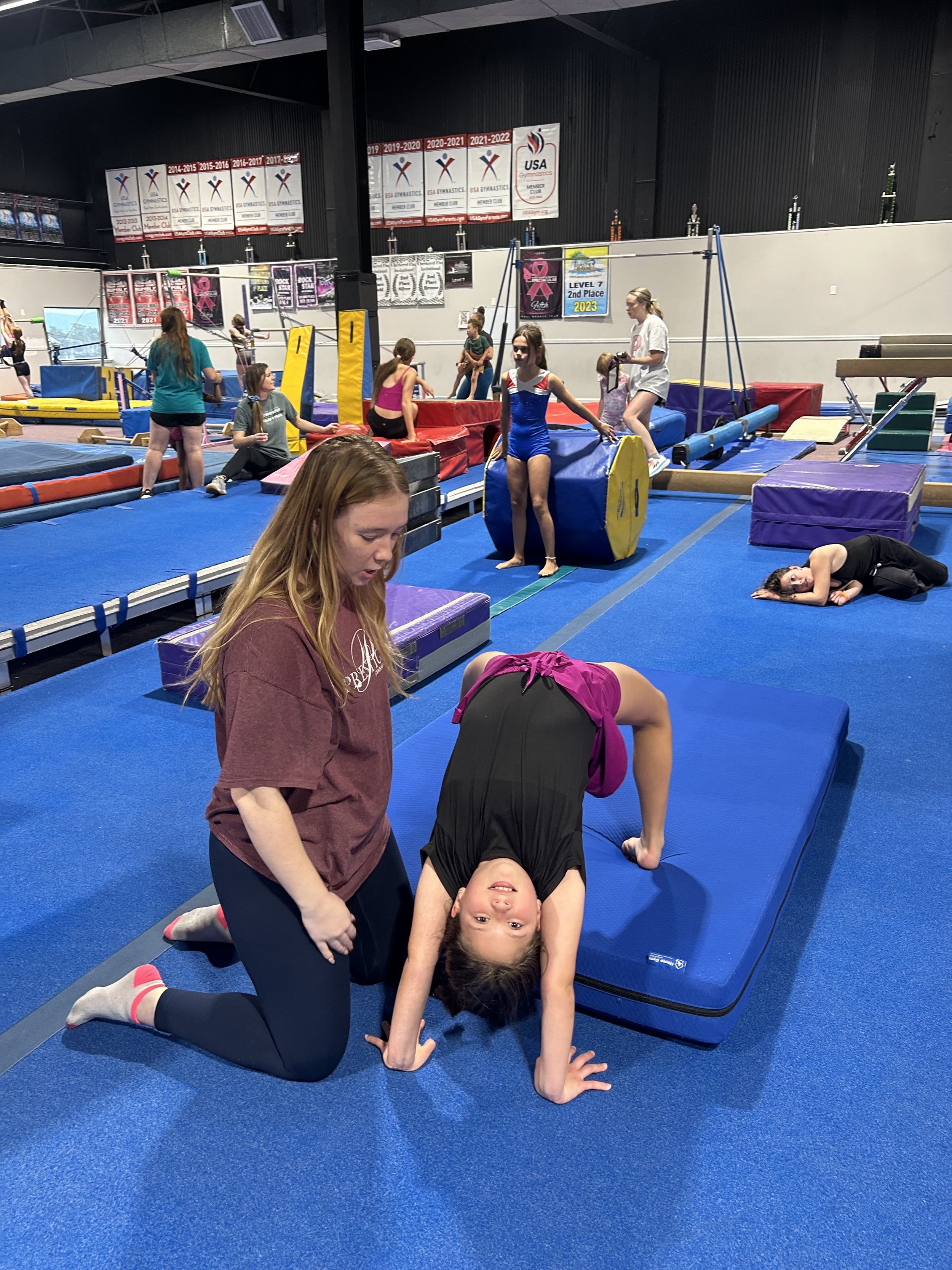 Four young girls in a gymnastics gym are forming a cheerleading lift; one girl is standing on the backs of two girls who are kneeling, with her arms raised in a cheer pose. An American flag hangs on the wall in the background.