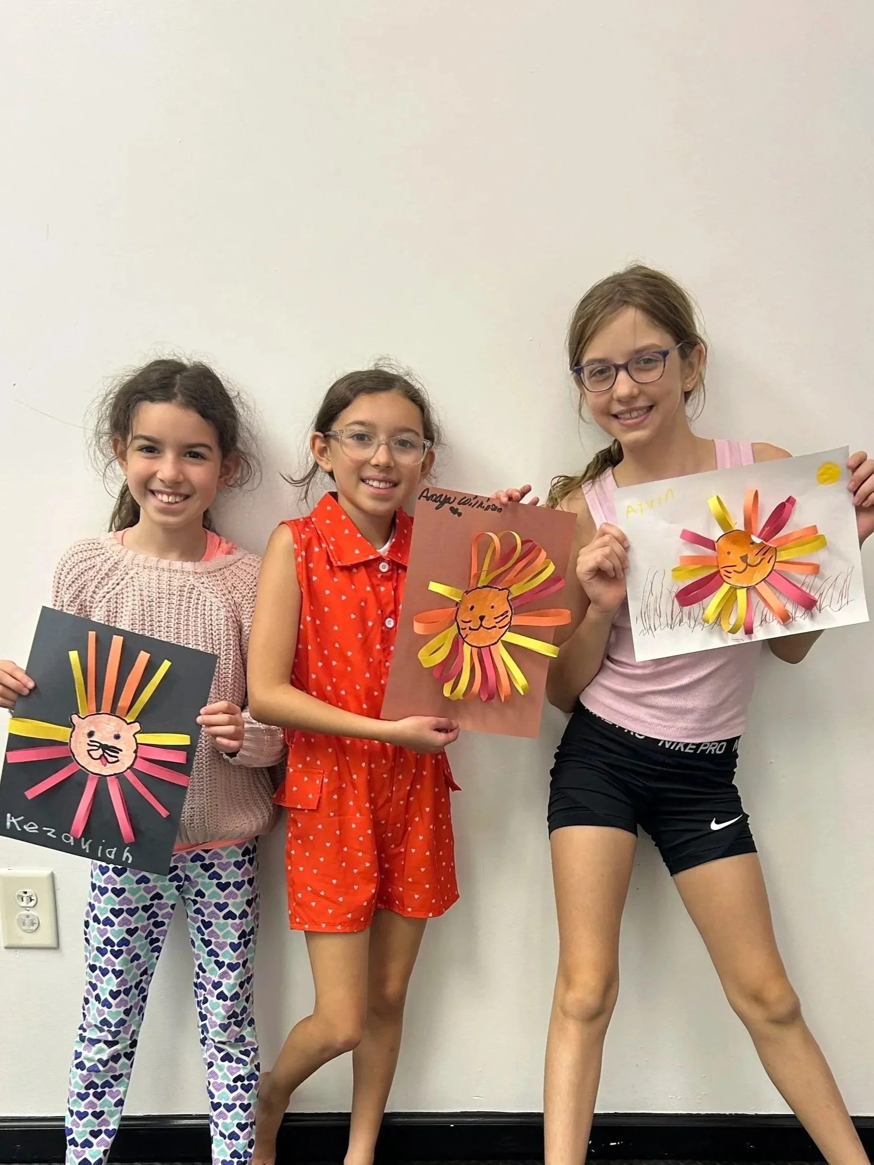 Children's hands drawing and coloring on paper at a table with markers, scissors, and paper crafts.