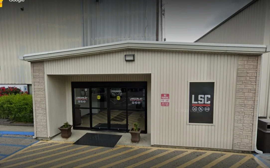 The entrance to a building with double glass doors, two potted plants outside, and a sign that reads 'No Parking Fire Lane.' The building is beige with vertical siding and has a window with the letters 'LSC' on it.