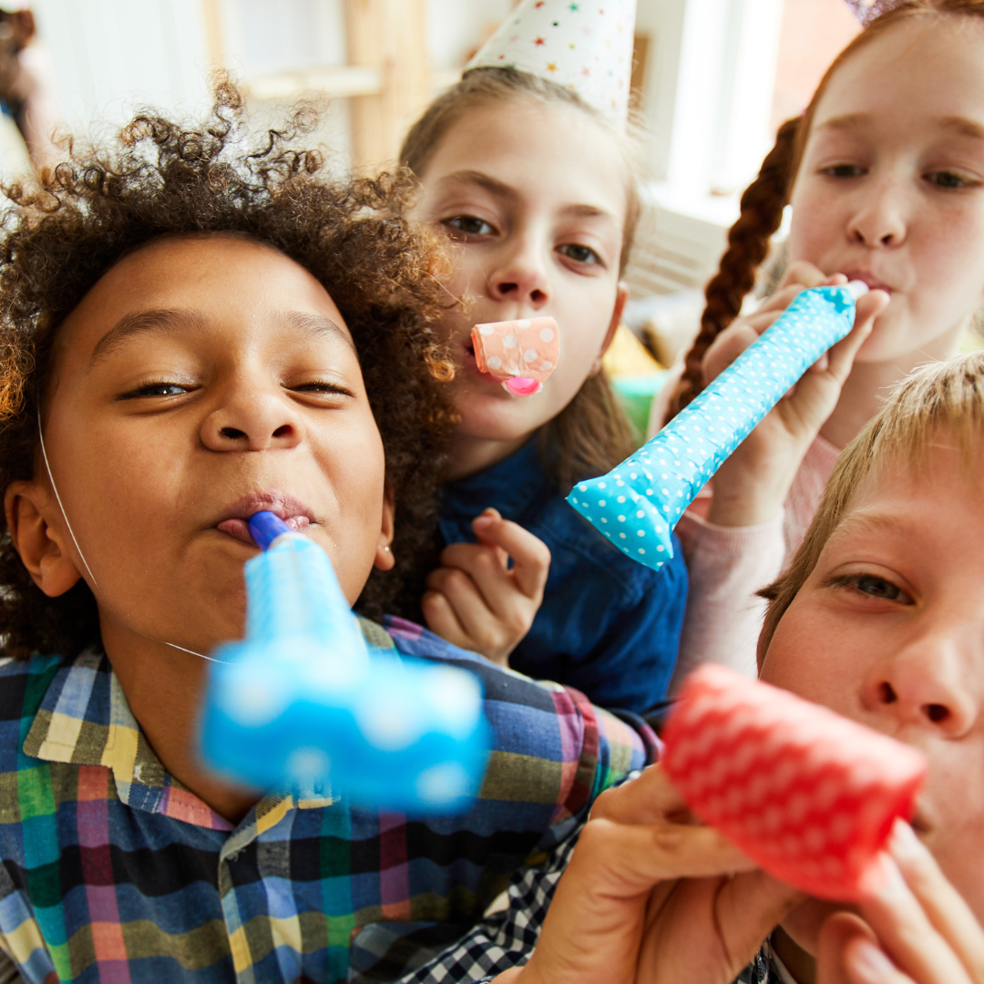 Group of children celebrating a birthday with party hats, blowing party horns, and wearing party accessories.