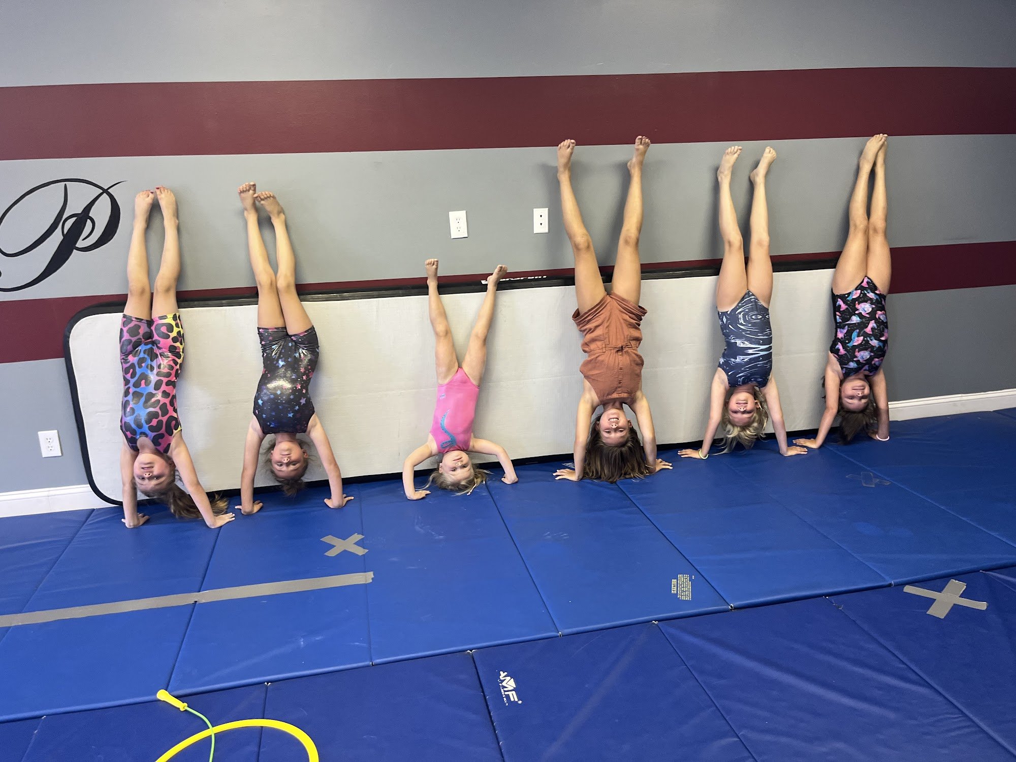 Six young girls performing handstands against a wall in a gymnastics or dance studio with a blue mat on the floor.