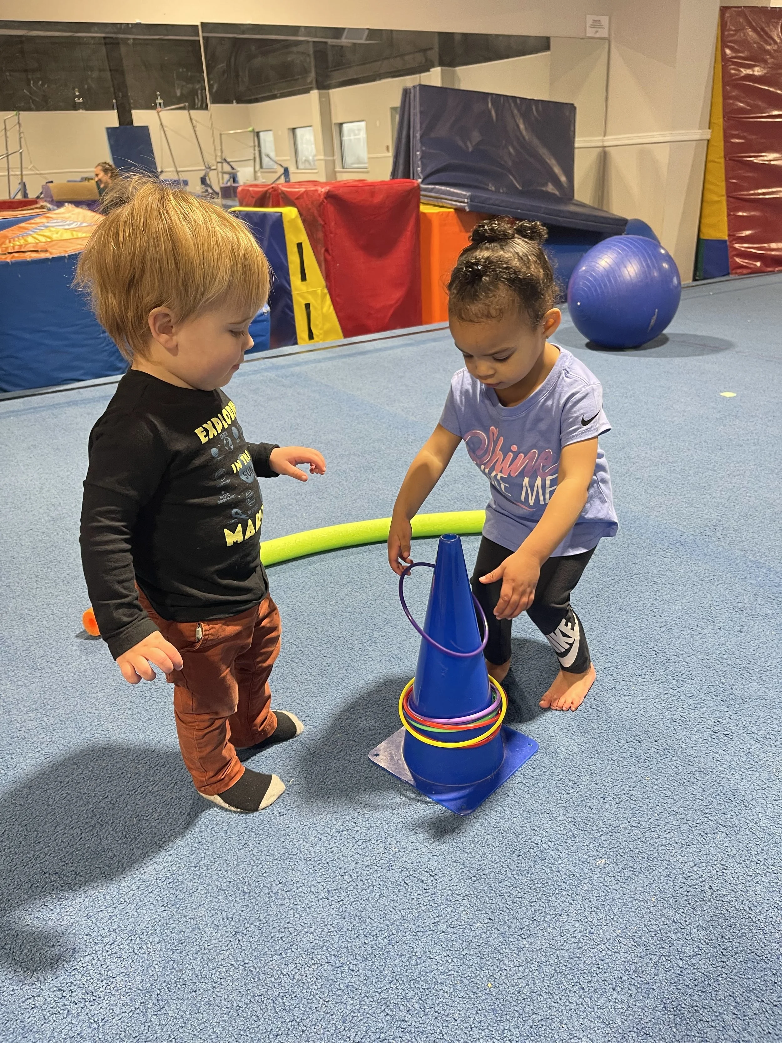 Two toddlers playing with colorful rings on a blue cone in an indoor play area.