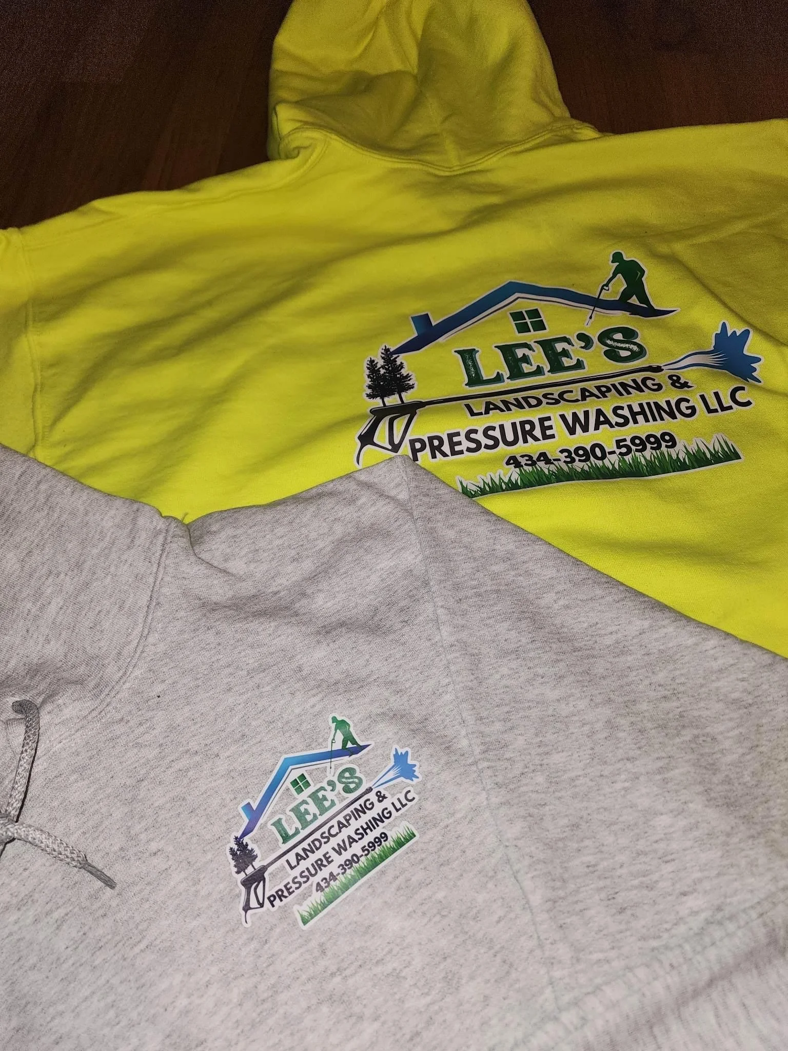 Yellow hoodie and gray sweatshirt both featuring a logo for Lee's Landscaping & Pressure Washing LLC, with a graphic of a person pressure washing a roof, trees, and a window, along with a phone number.