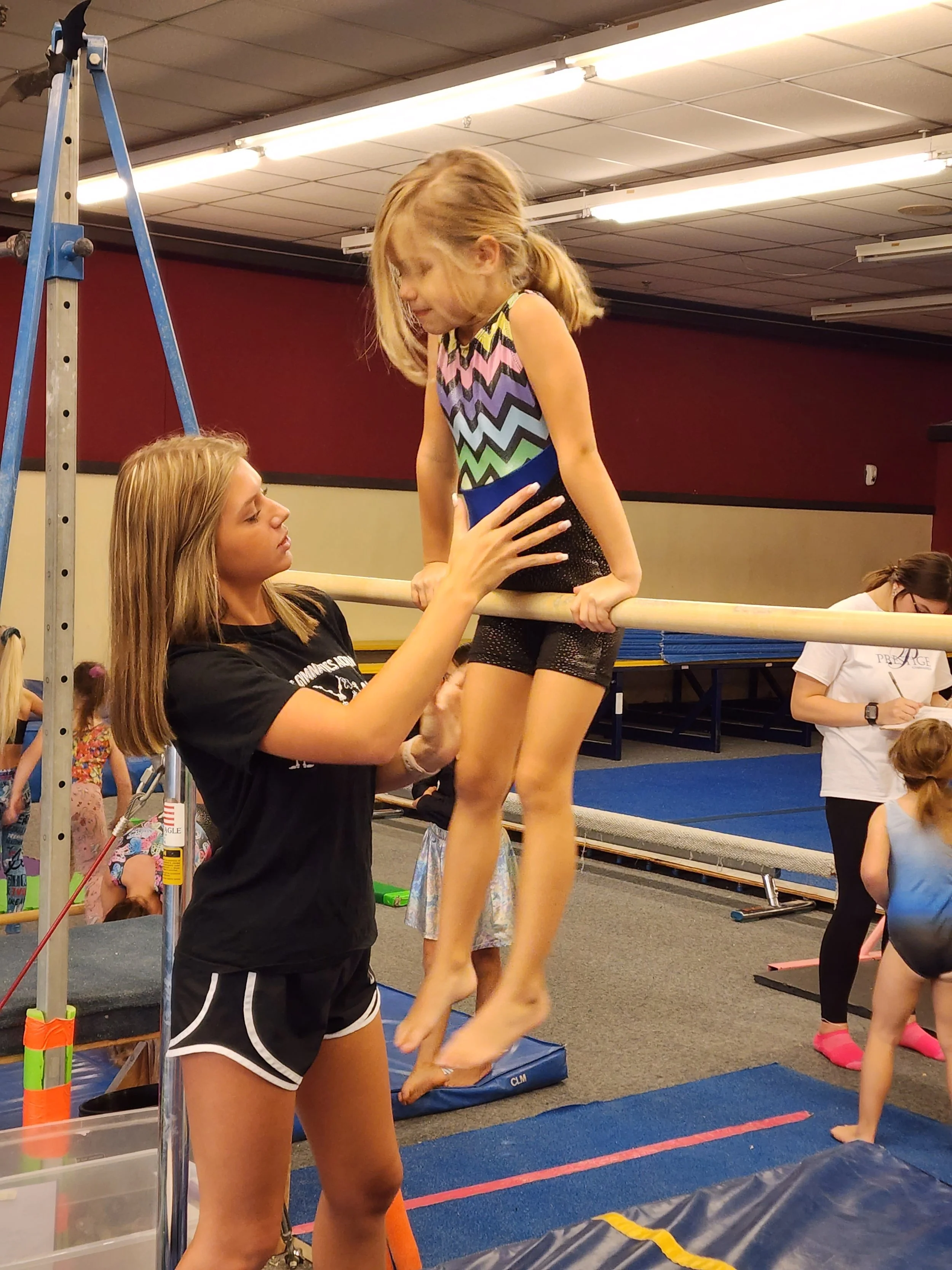 A girl practicing uneven bars at an indoor gymnastics facility, with an instructor assisting her.