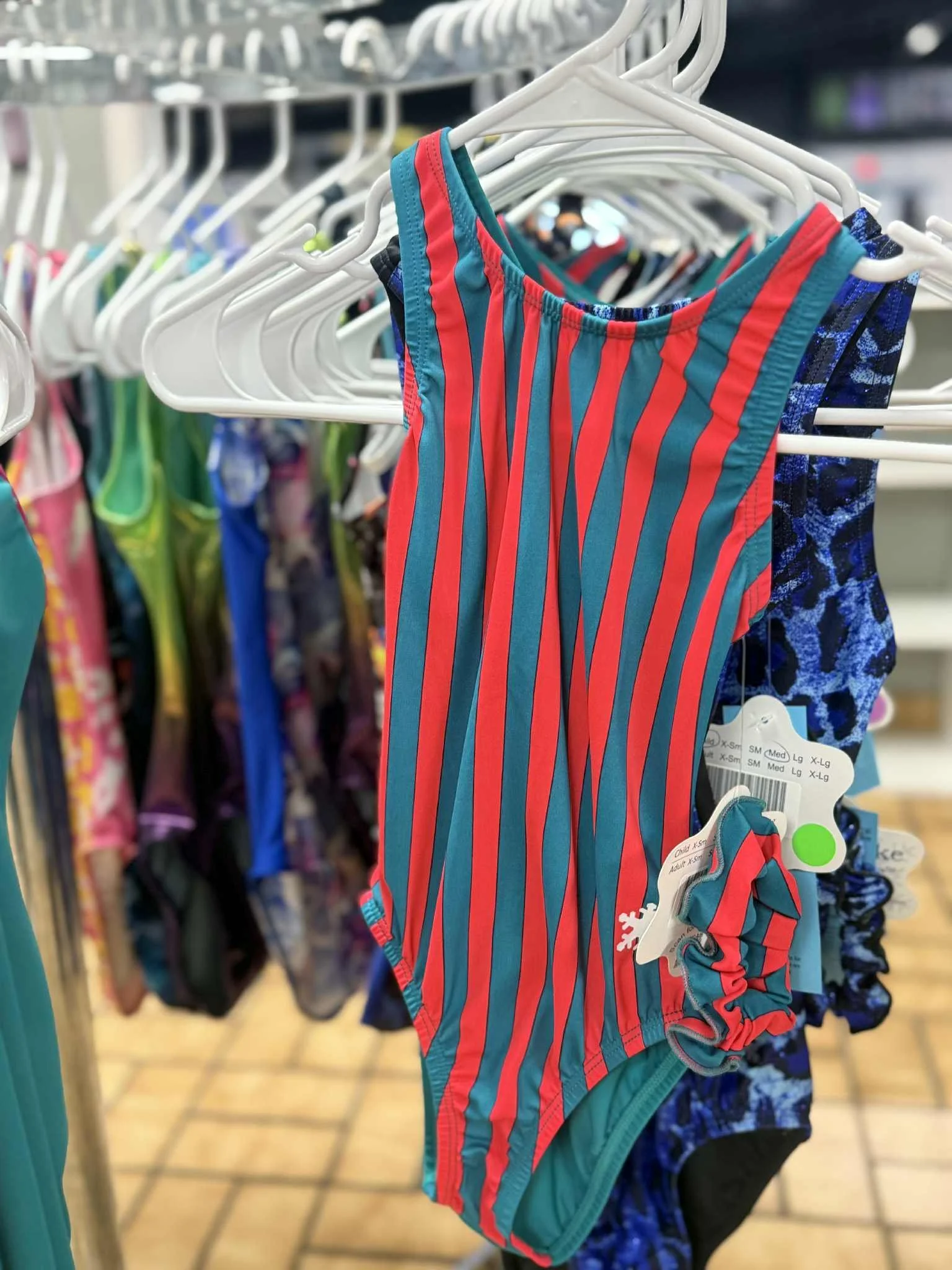 Various athletic clothing, including colorful swimsuits, a green 'gymnast' t-shirt, and shorts, displayed on hangers in a retail store.