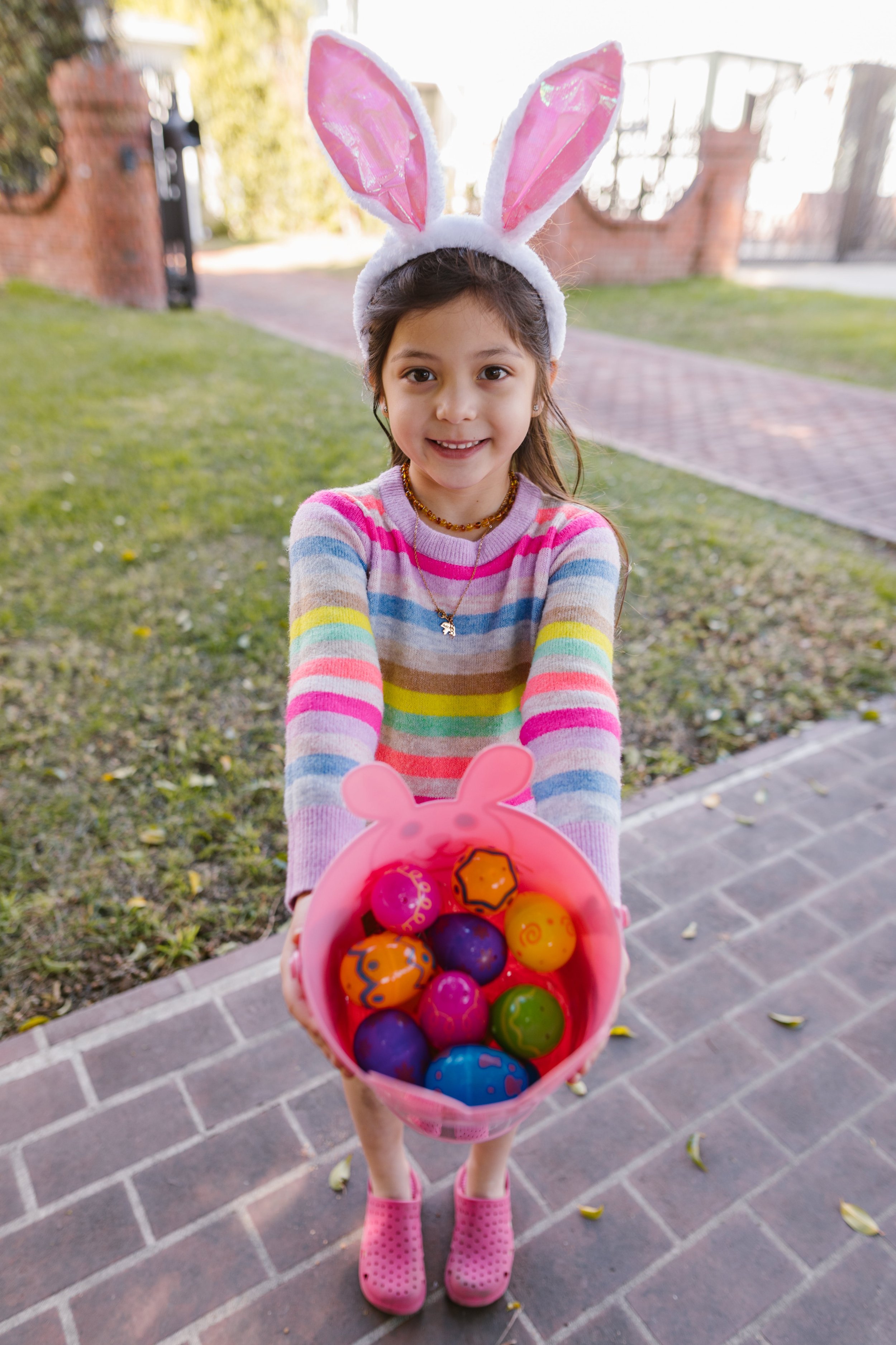 A young girl wearing a rabbit ear headband and a colorful striped sweater is smiling and holding a pink bunny-shaped basket filled with decorated plastic Easter eggs. She is standing on a brick sidewalk outdoors with grass, trees, and a brick wall in the background.