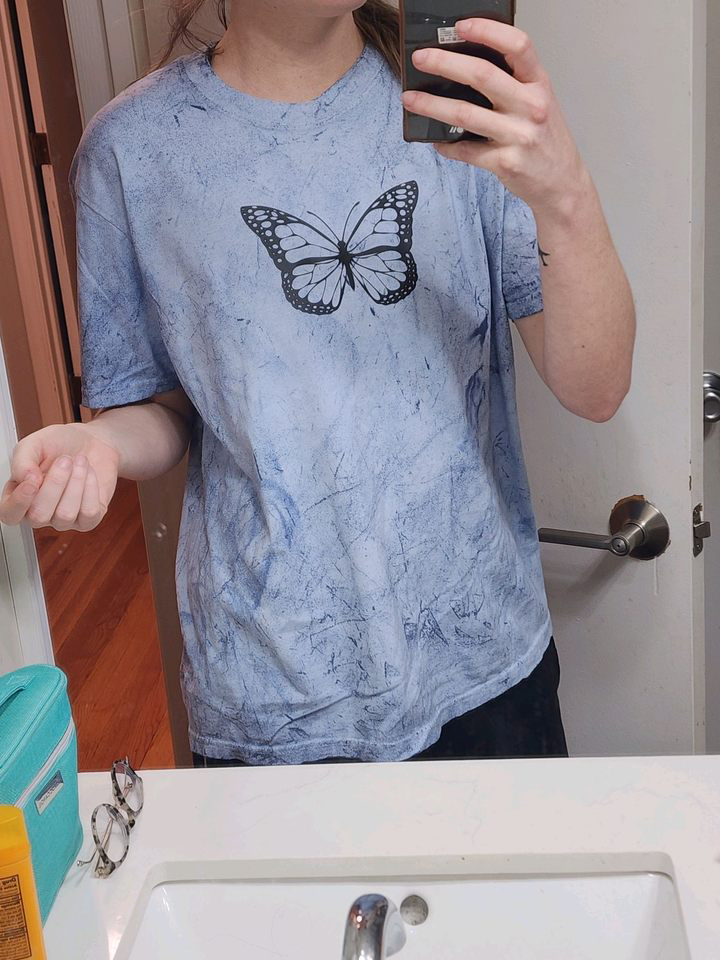 Person taking a mirror selfie wearing a light blue tie-dye t-shirt with a butterfly graphic on the front, in a bathroom with a white sink, glasses, and toiletries visible.