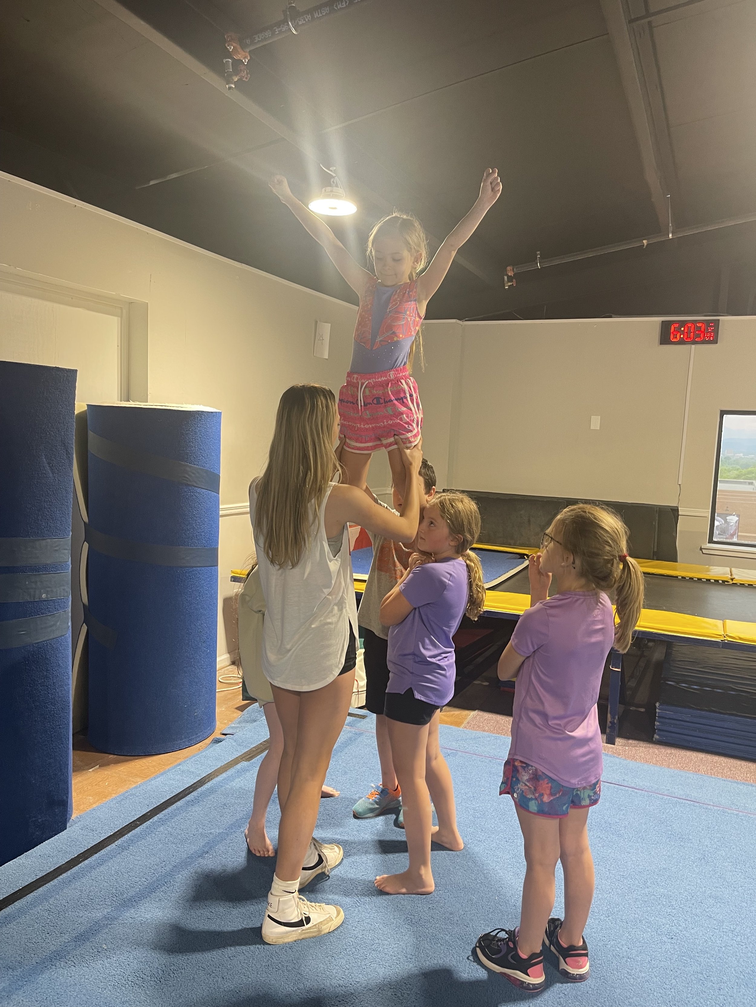 Children practicing cheerleading or gymnastics with a girl standing on an adult's hands in an indoor gym.