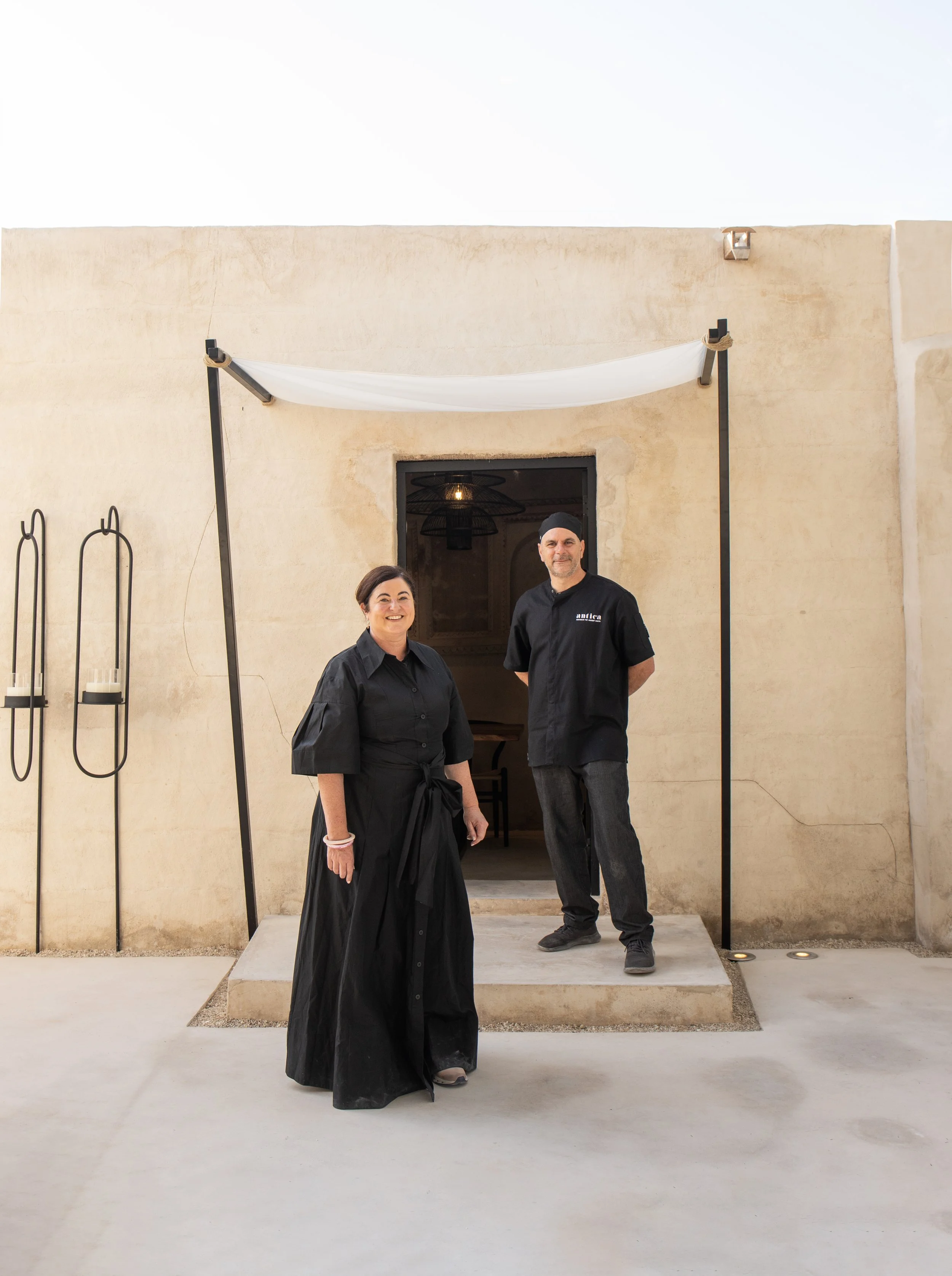 Kelly and Paolo Picarazzi dressed in black and standing outside a sand coloured building with a white sail cloth above them. In Al Jazeeraa Al Hamra Heritage Village of Ras al Khaimah in the UAE